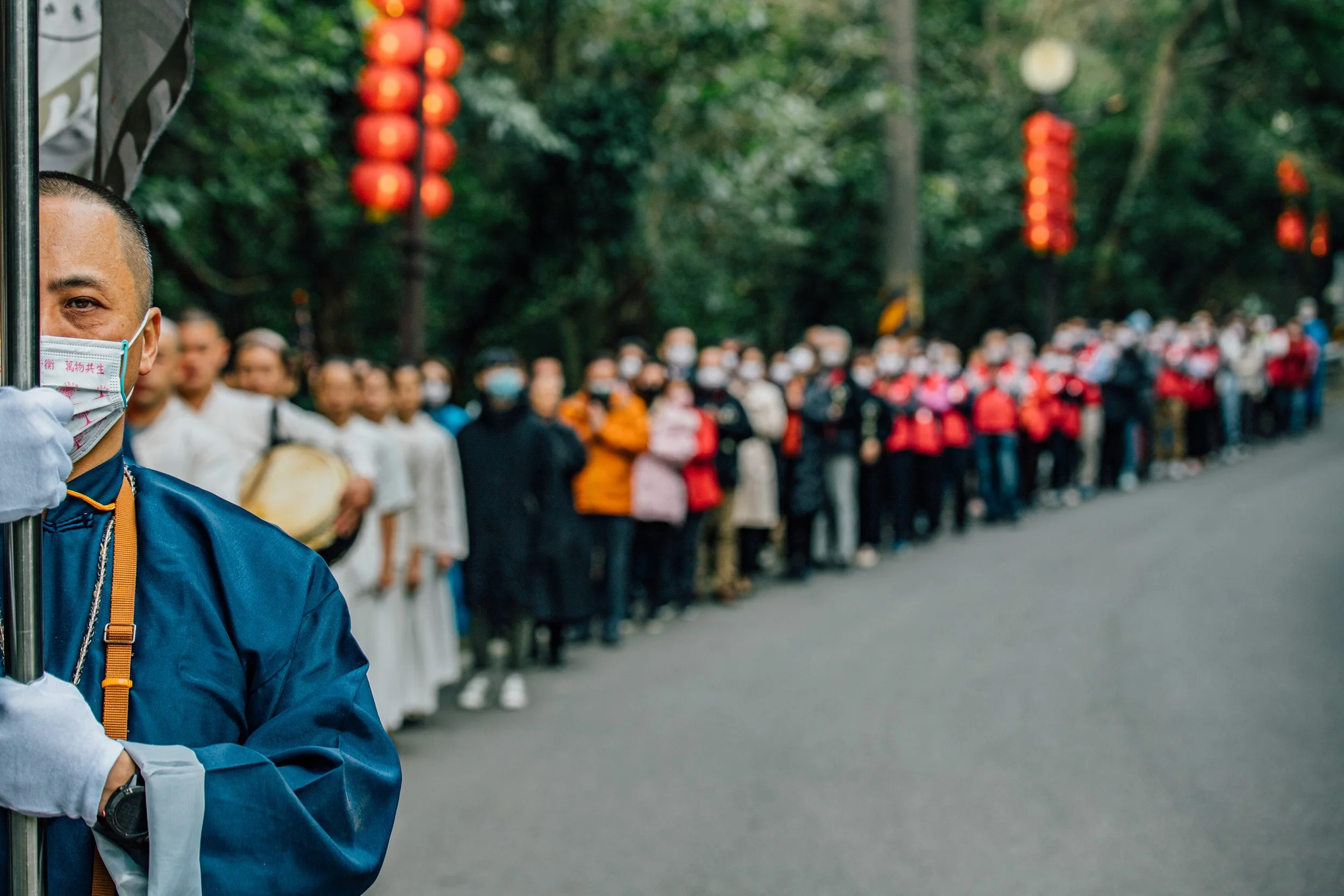 Close-up of a man wearing a face mask and a blue jacket, standing on the side of a street with a crowd of people in the background, all wearing masks and dressed warmly. The scene appears to be an outdoor gathering or event, with red lanterns hanging
