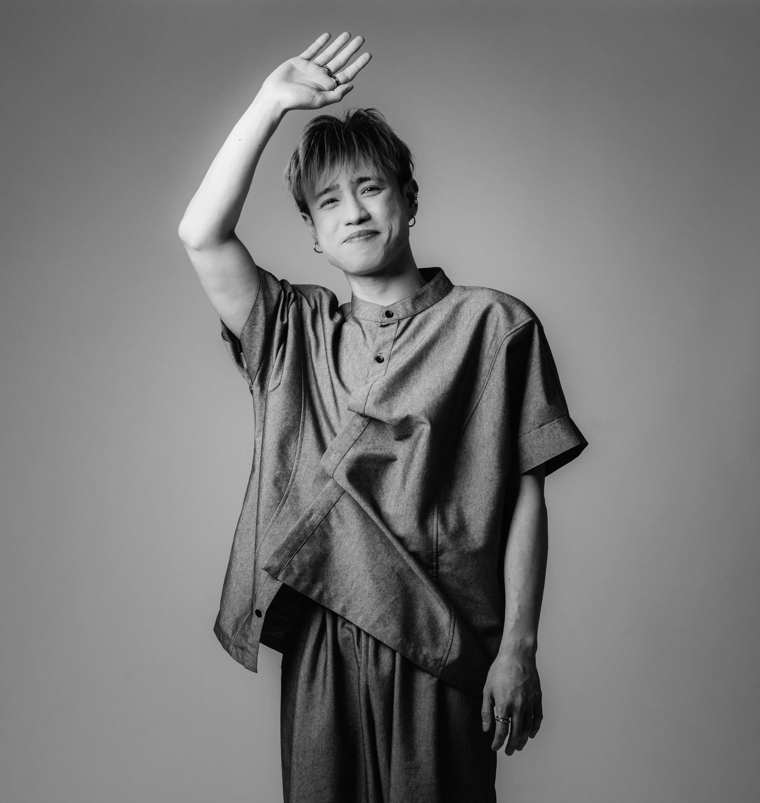 Black and white photo of a young person with short hair, wearing a silky shirt, raising their right hand with a few rings, and smiling with their lips closed.