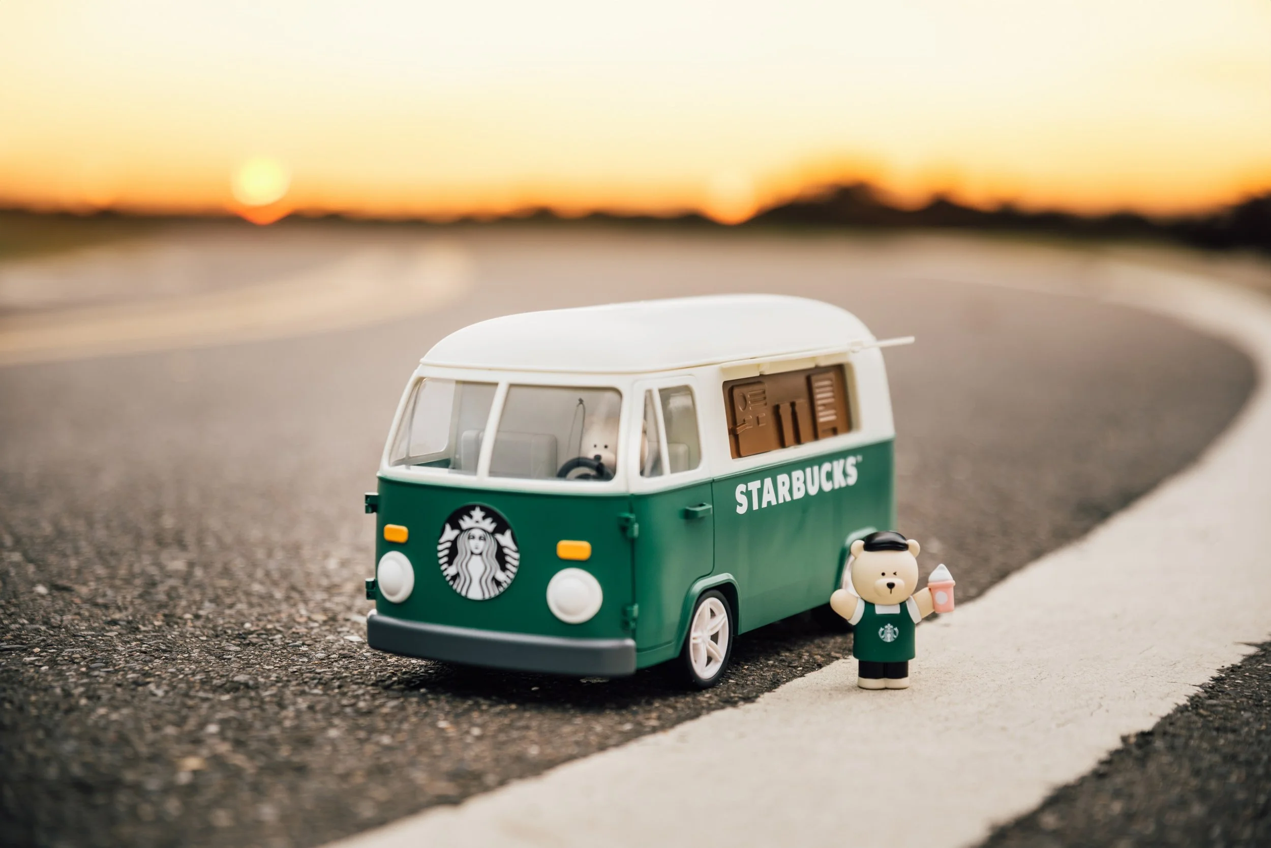 A small toy van resembling a Vancouver-style bus with Starbucks branding, alongside a small bear figure in Starbucks attire, on a road with a blurred sunset in the background.