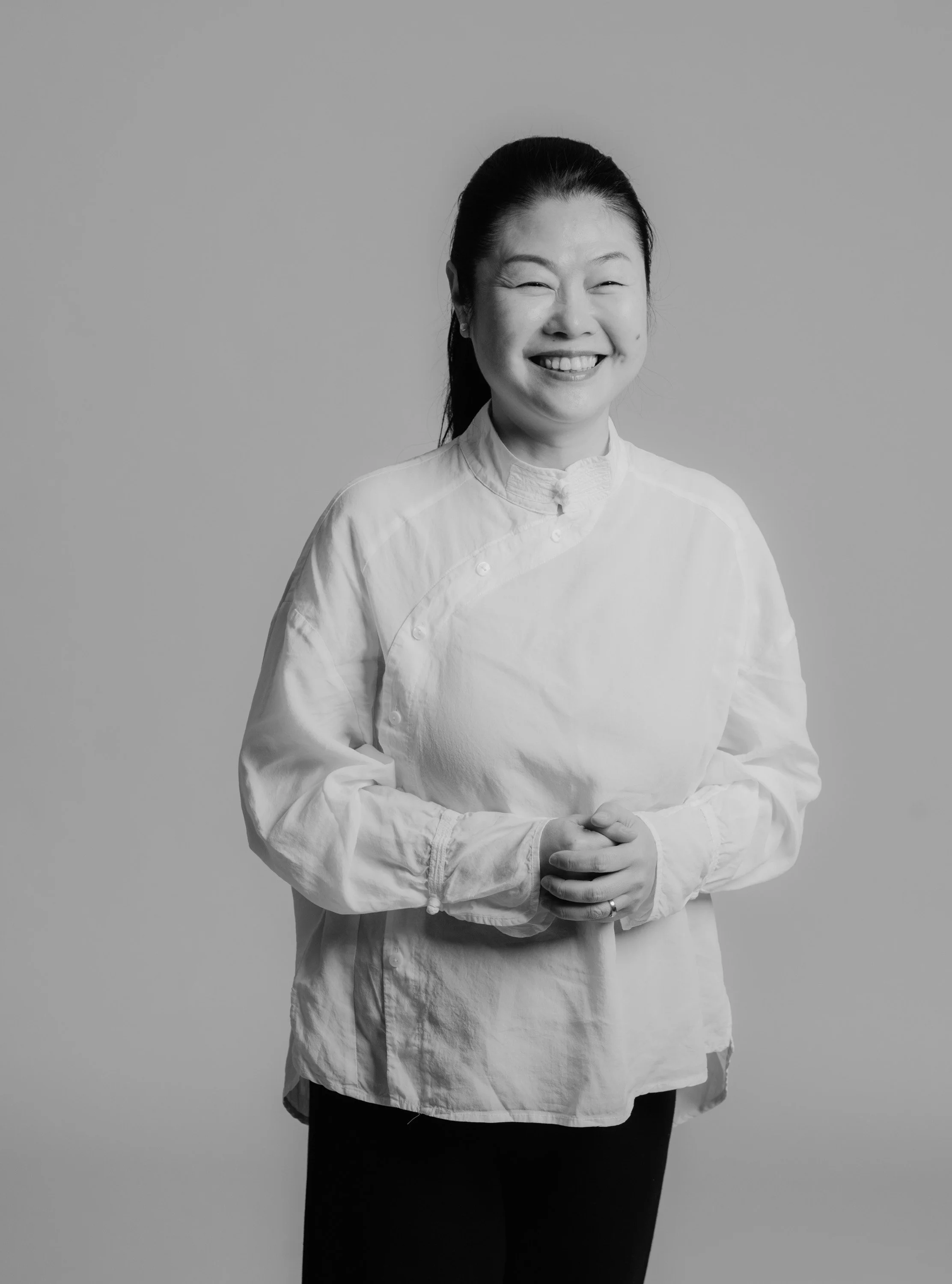 A woman wearing a chef's jacket standing against a plain background, smiling with her hands clasped in front of her.