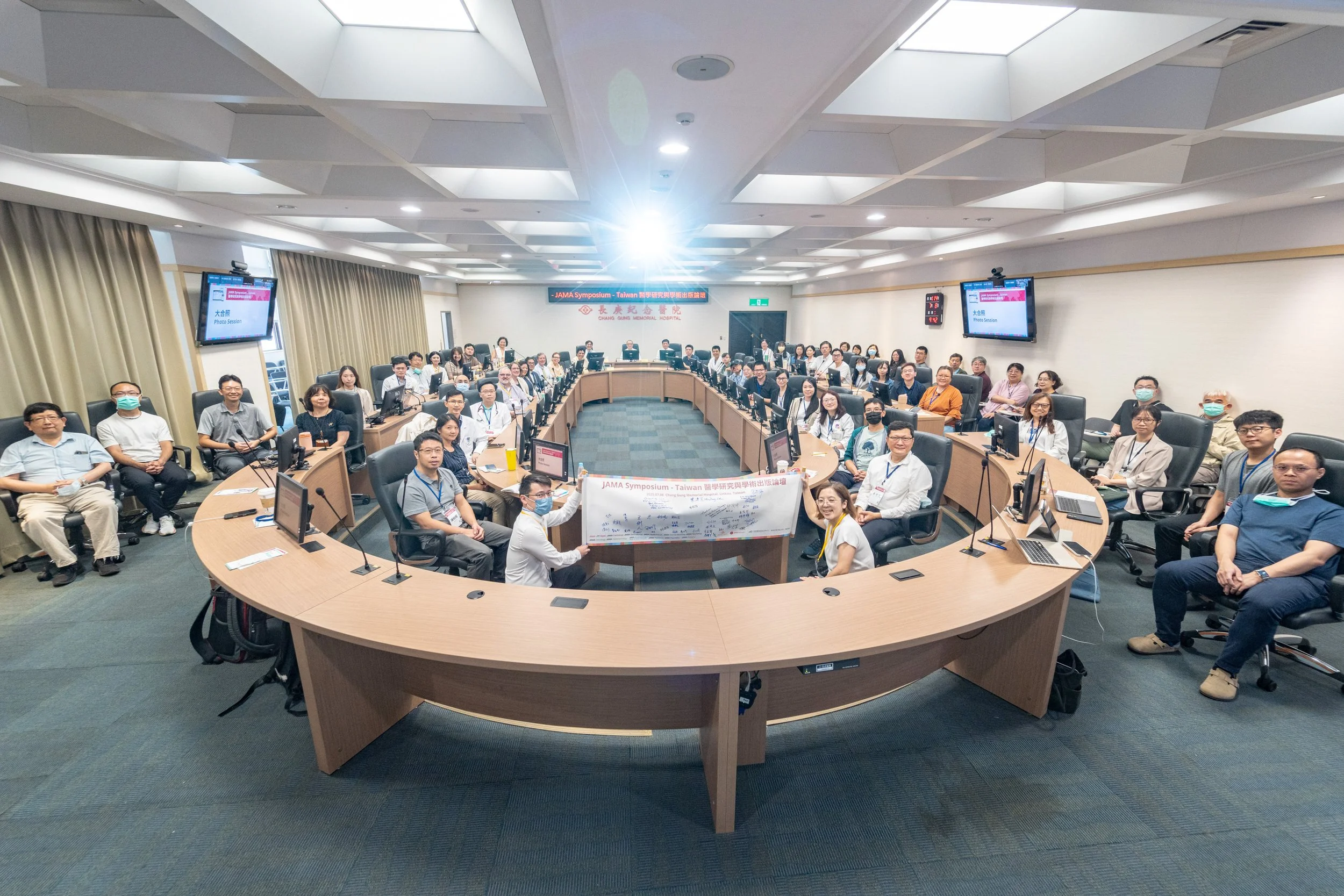 A large conference room filled with people sitting around a U-shaped arrangement of tables, facing the camera. The room has monitors, laptops, and microphones on the tables. A banner on the wall indicates the event is the JAMA Symposium in Taiwan at 