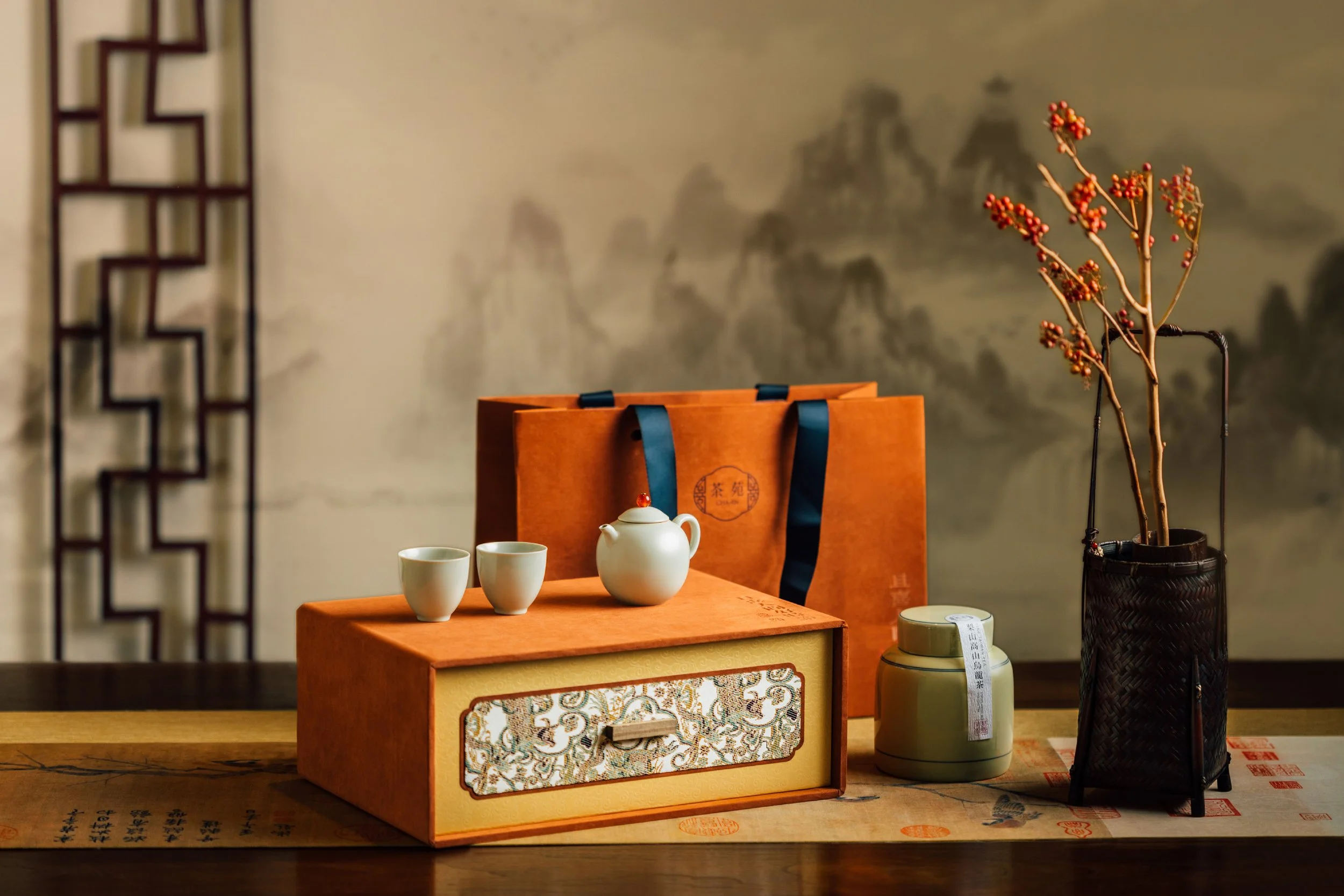 A tabletop display featuring a white teapot with a lid, two small matching teacups, a decorative box with traditional Asian design, a paper bag with Asian characters, a white jar with a ribbon, and a small decorative plant in a black basket, all set 