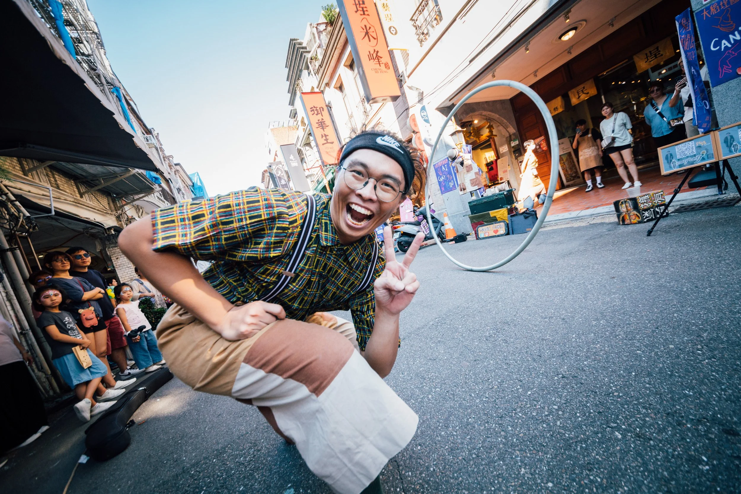 A young man smiling and making a peace sign with his fingers in the street, with a crowd of people watching behind him and storefronts with colorful signs in an urban shopping district.
