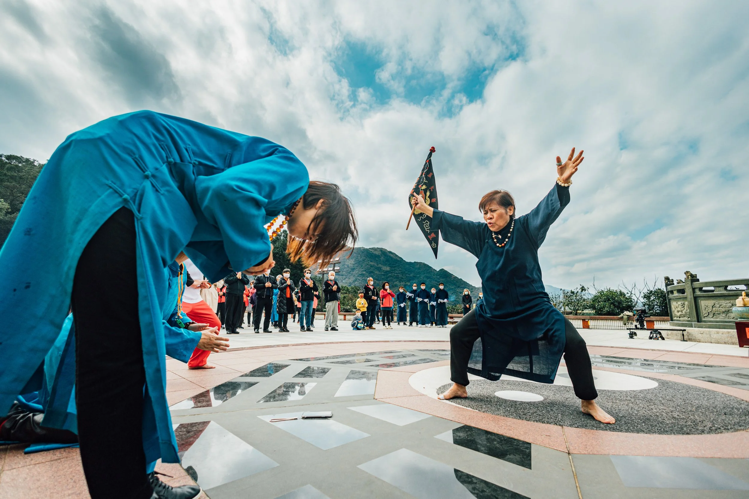 A person dressed in traditional attire performing a martial arts move in front of an audience, some of whom are bowing, outdoors with cloudy sky and mountains in the background.