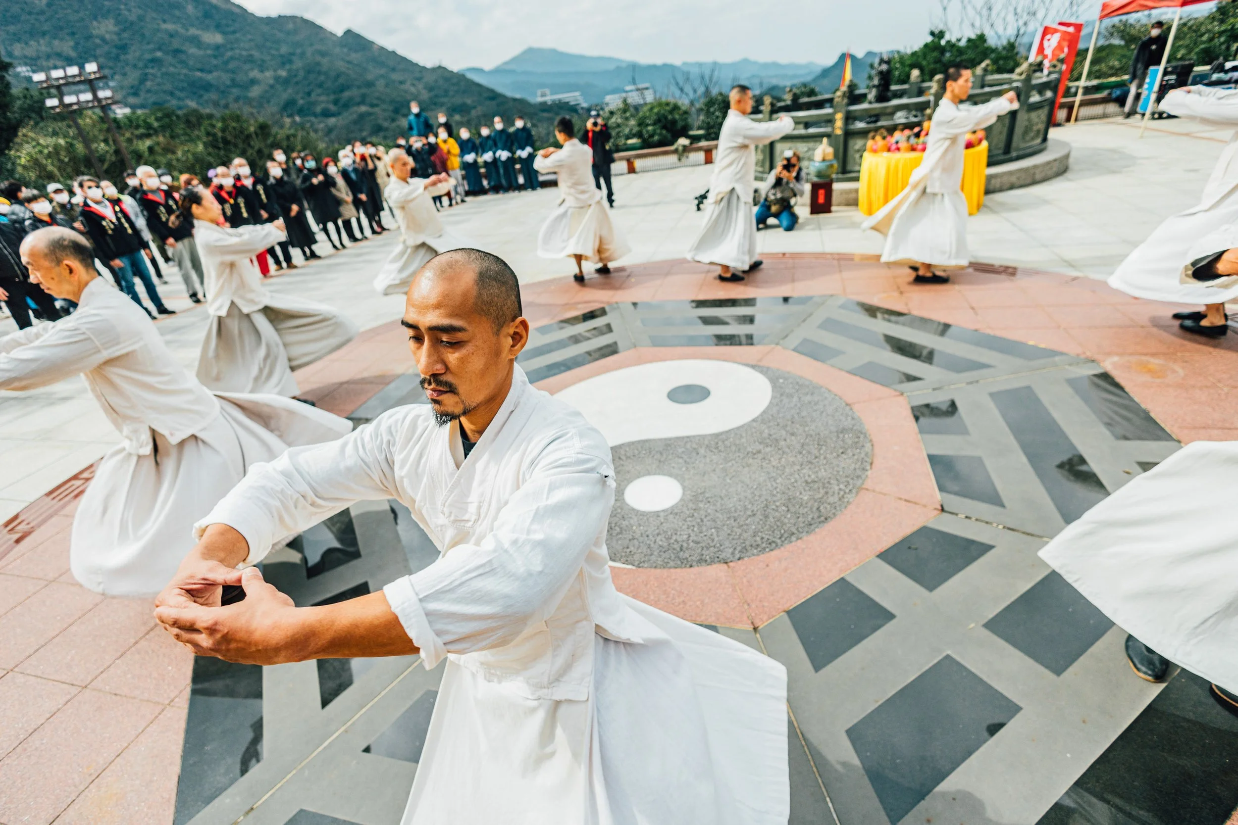 A group of people dressed in traditional martial arts uniforms performing a synchronized martial arts routine outdoors on a circular pagoda-style platform with a yin-yang symbol. In the background, there is an audience and mountains under an overcast