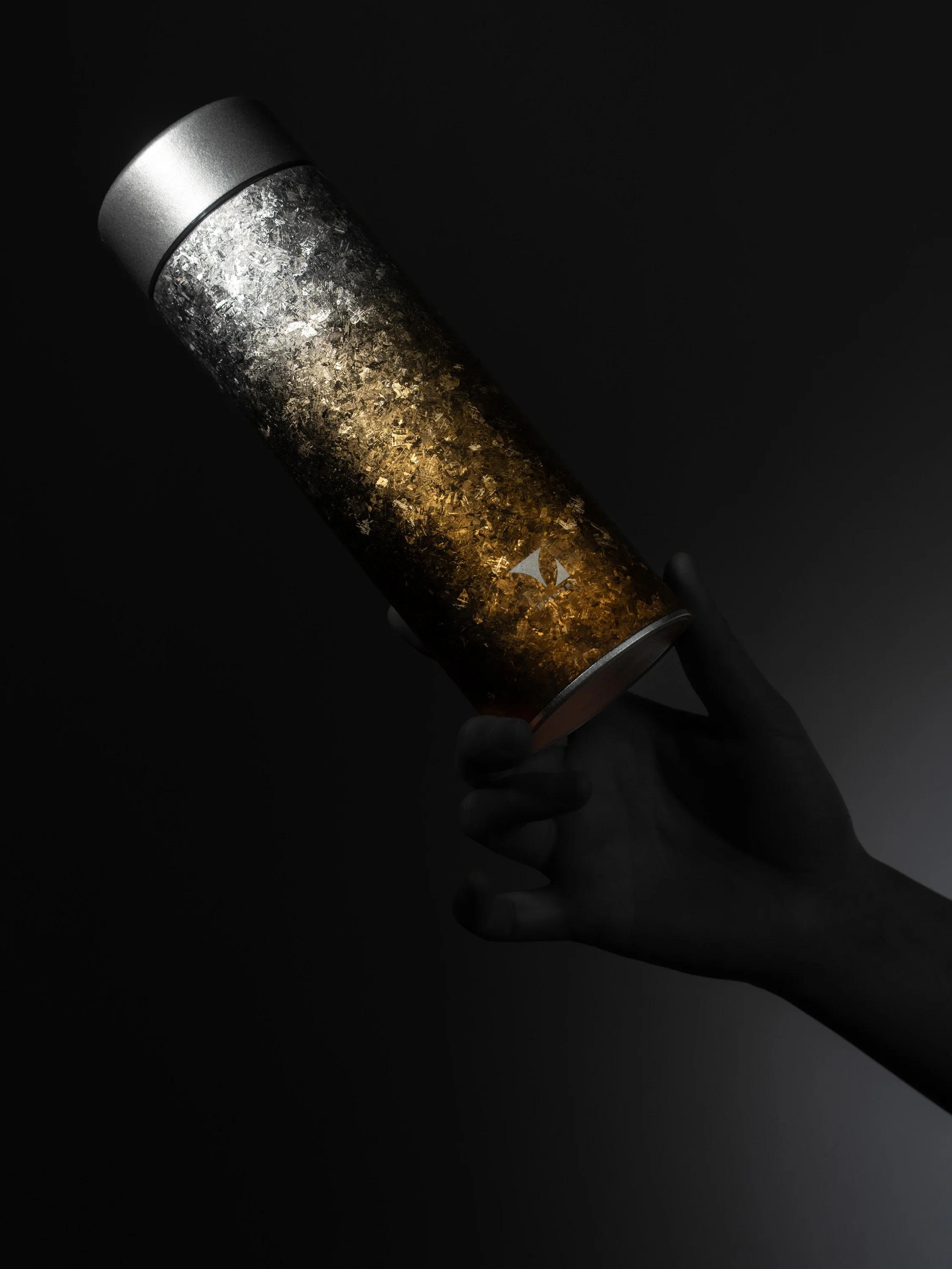 A hand holding a black tumbler with a metallic lid, filled with a dark beverage and gold and silver metallic flakes at the bottom.