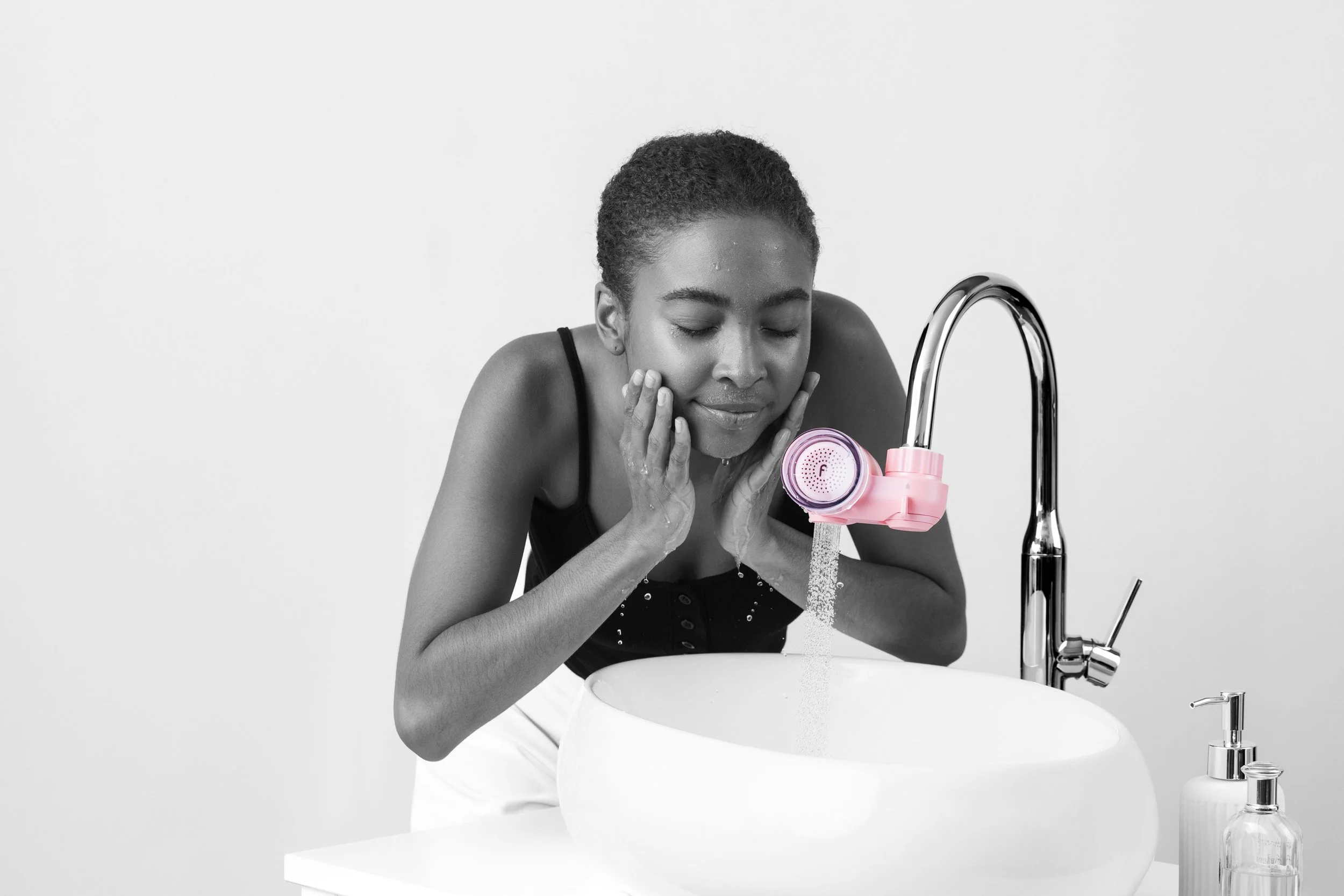 A woman washing her face at a bathroom sink with a pink facial cleansing device, water running from the faucet, and a soap dispenser in the foreground.