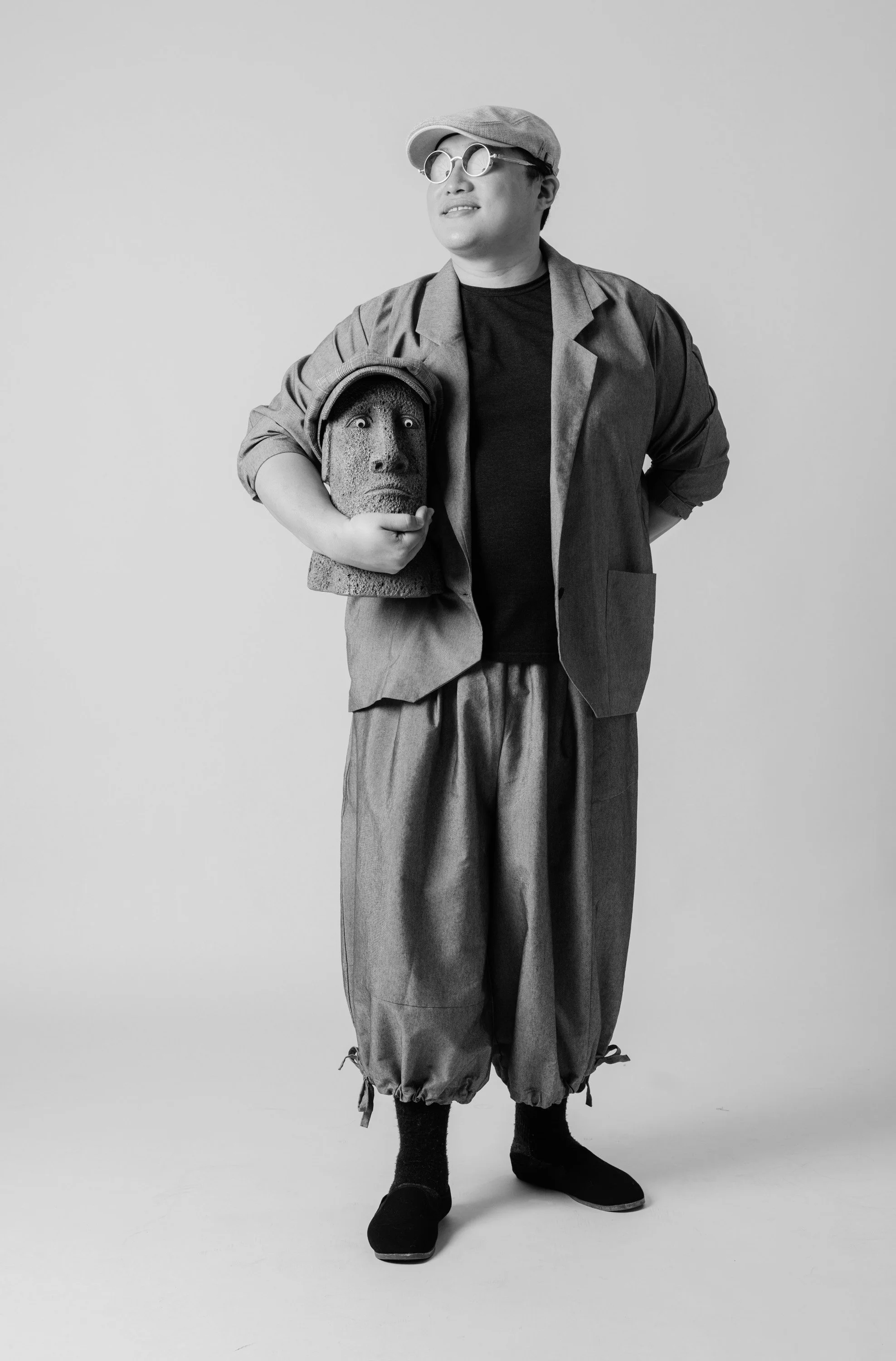 A person dressed in layered, loose-fitting clothing, including a cap, sunglasses, blazer, baggy pants, and boots, standing in front of a plain background, holding a carved stone sculpture with a surprised facial expression.