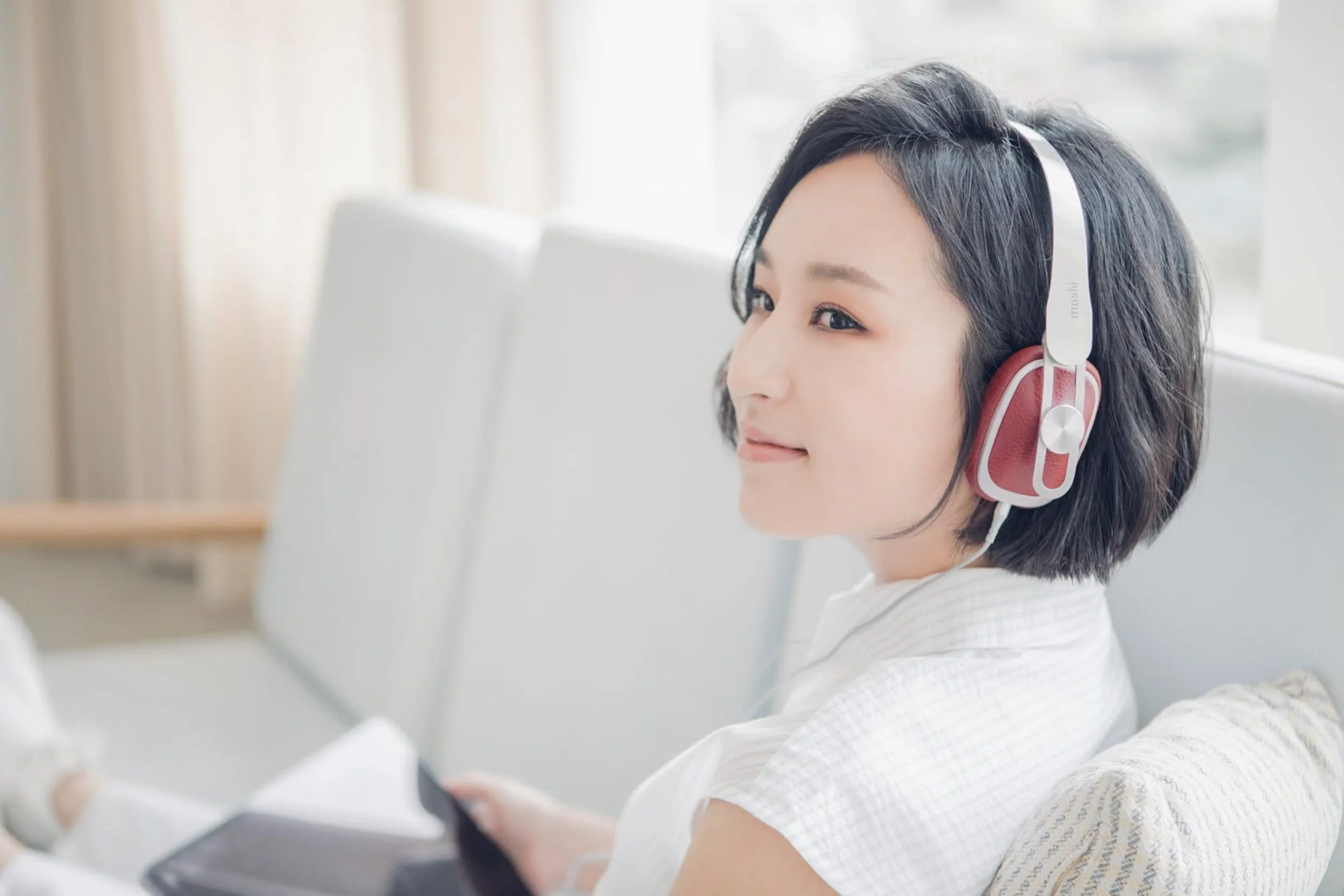 A young woman with short black hair wearing a white shirt and headphones with red ear pads, sitting at a desk, looking at a computer screen in a bright room with natural light.