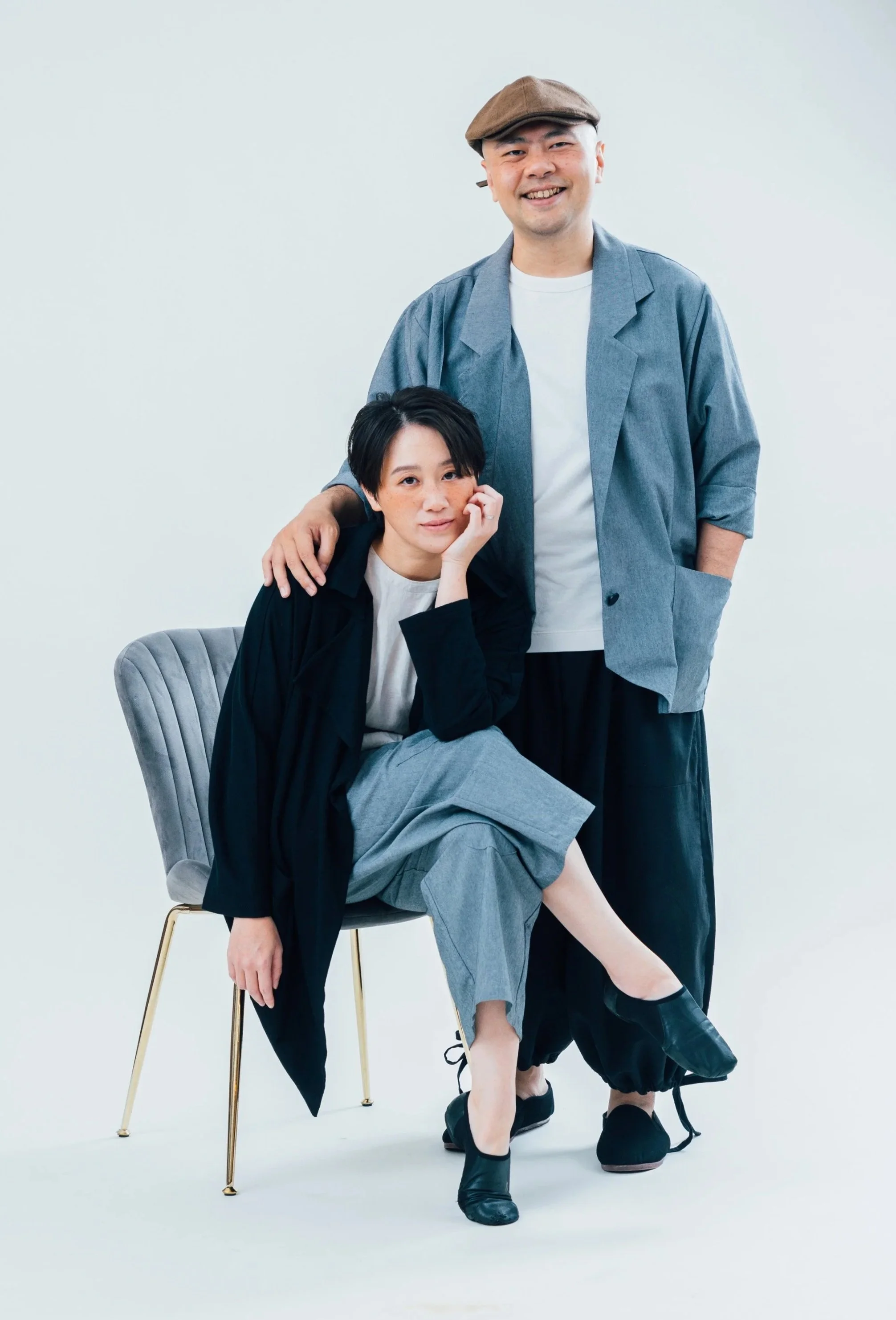 A man and woman pose together against a plain white background. The woman is sitting on a chair, resting her head on her hand, with short black hair and wearing a black jacket and gray pants. The man stands behind her with his hand on her shoulder, smiling, wearing a brown cap, gray jacket, white shirt, and black pants.
