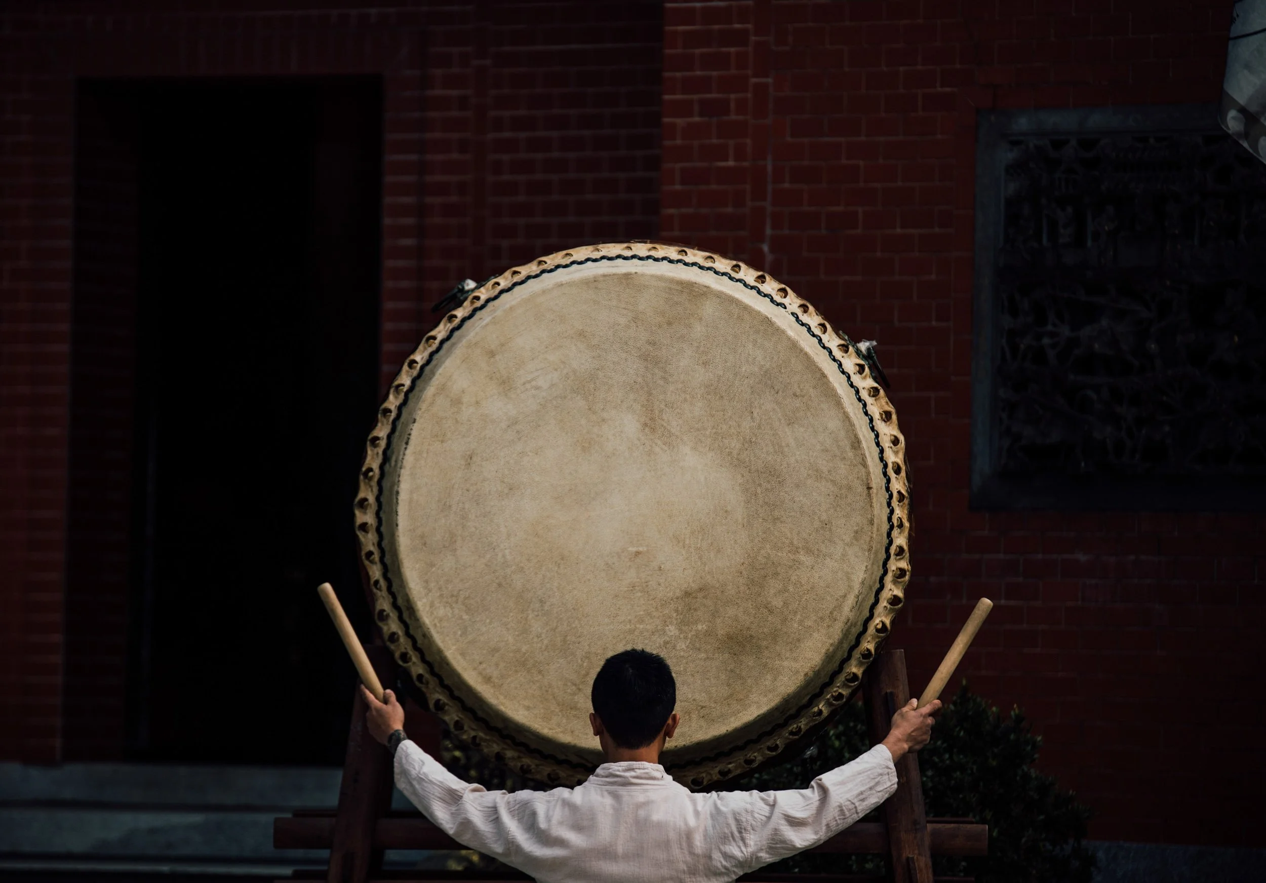 Person in traditional white attire playing a large drum. The drum is mounted on a stand, and the person is holding drumsticks. The background features a brick wall.
