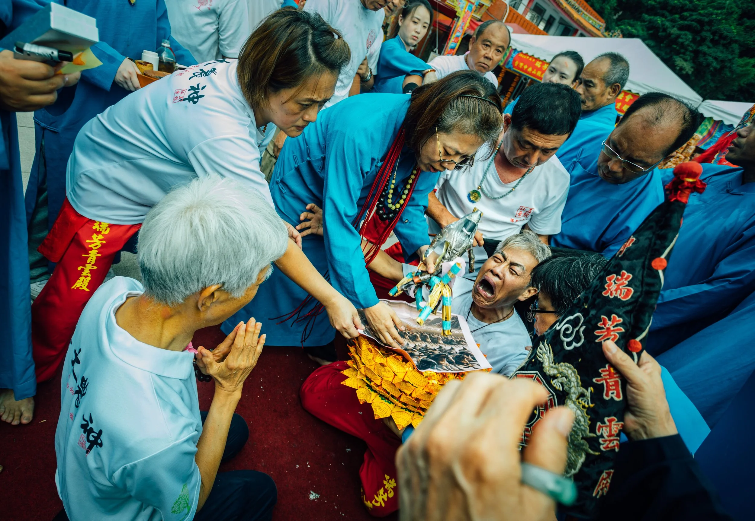 Group of people gathered outdoors during a cultural event, with some participants dressed in traditional attire and engaging in an activity involving a portrait and festive decorations.
