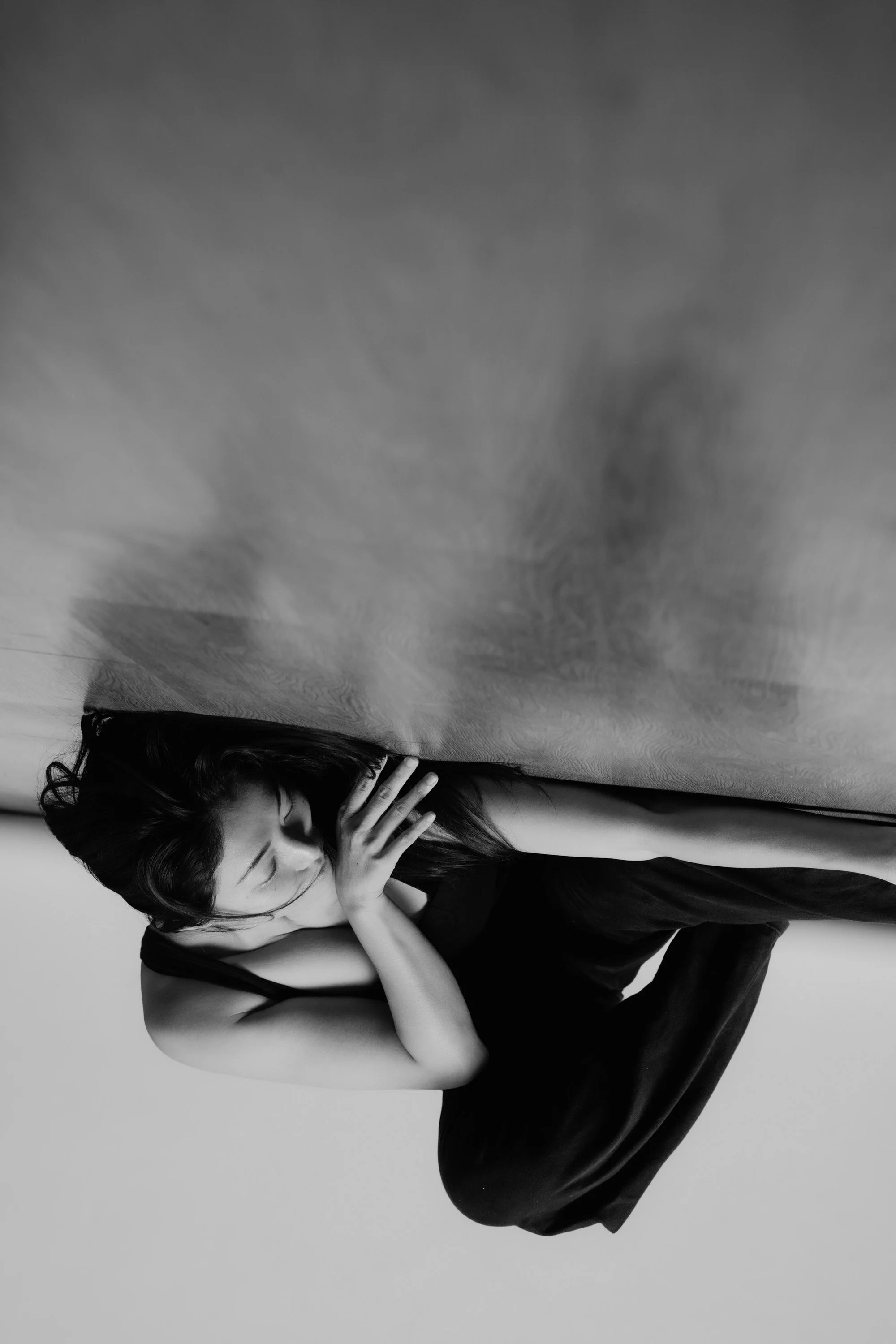A woman with black hair, a black top, and black pants is sitting on the floor with her head resting on her knees, and her hands covering her face. The photo is in black and white.