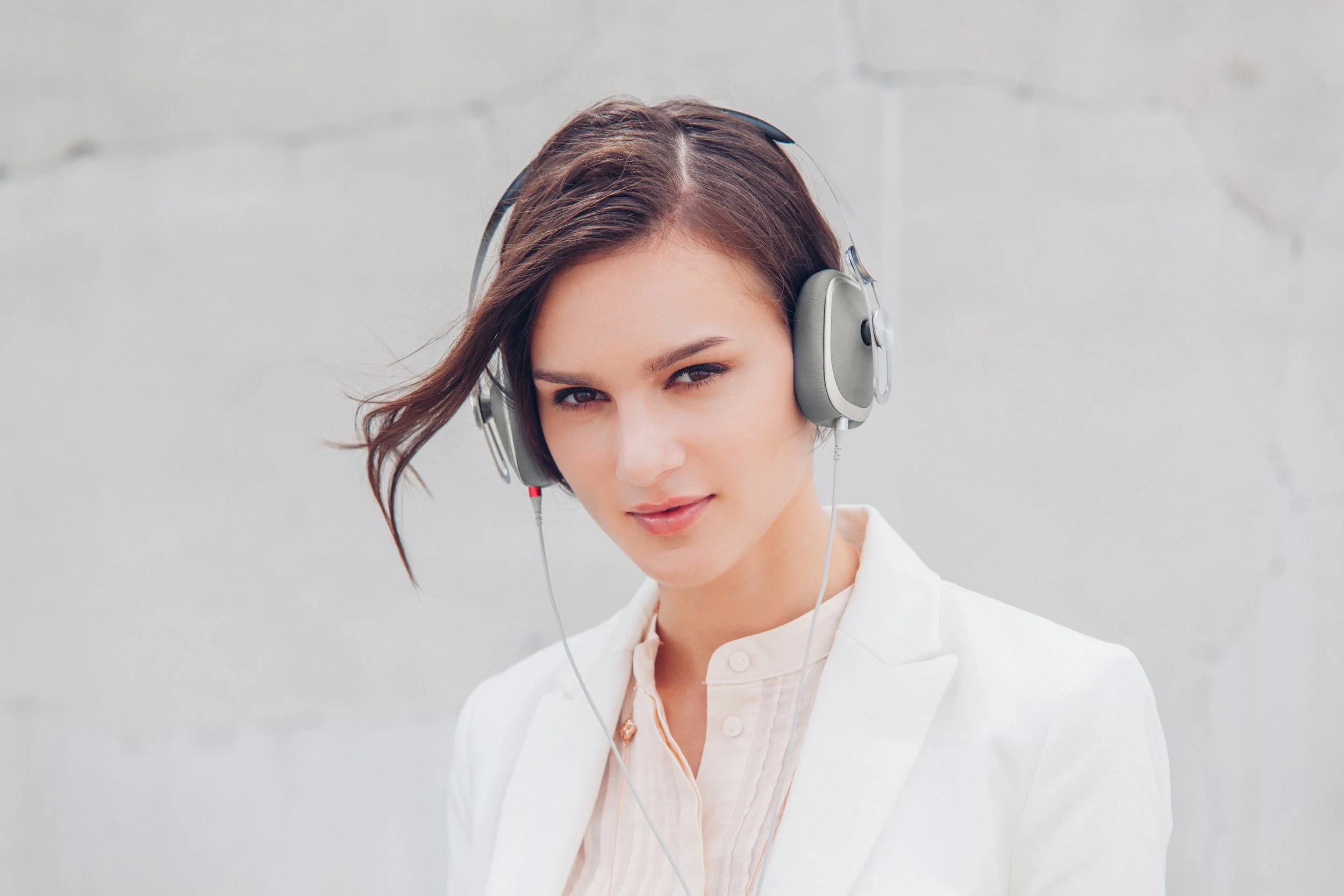 A woman with brown hair wearing headphones and a light-colored blazer, standing in front of a plain light gray background.
