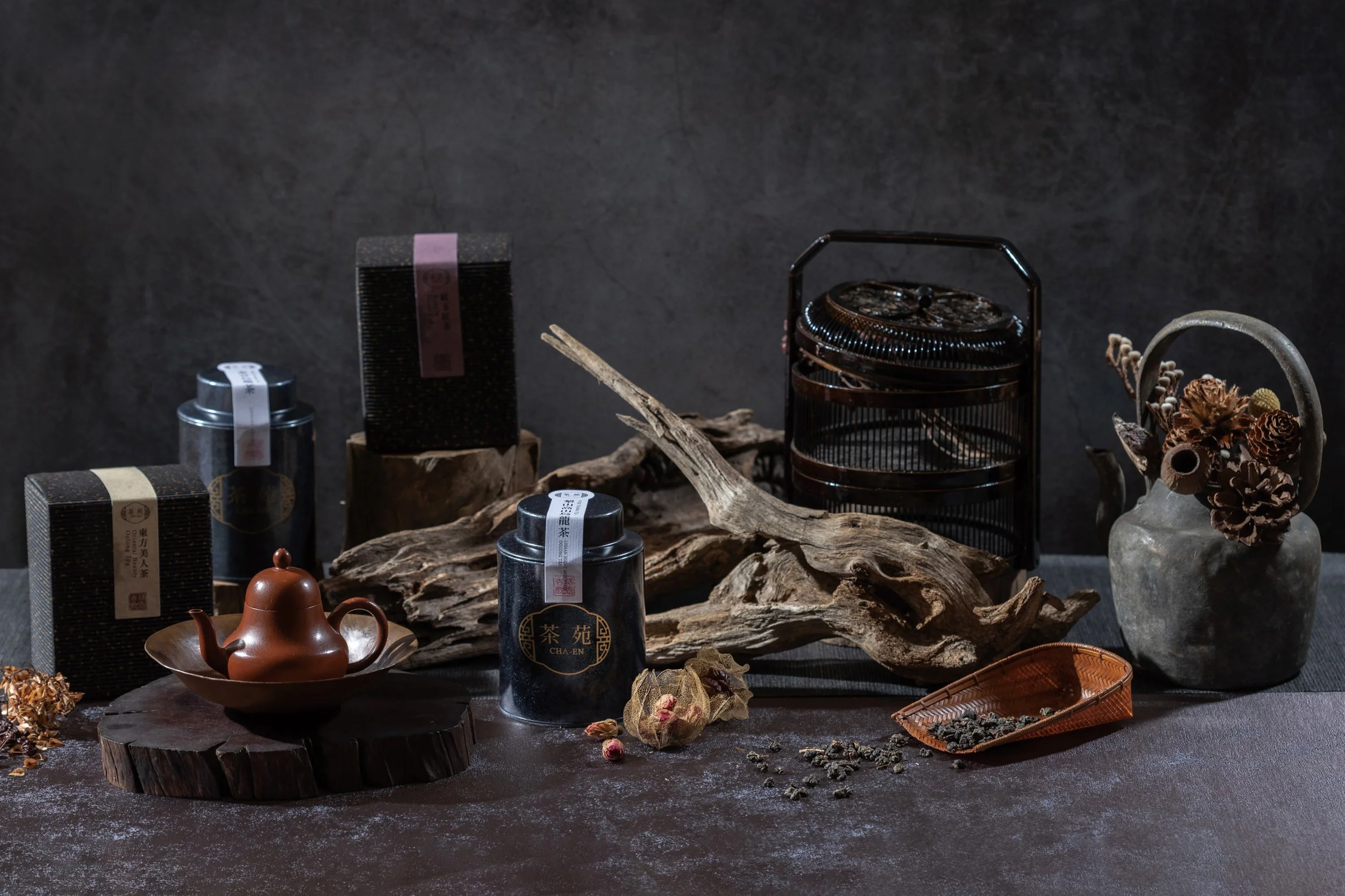 A still life arrangement of tea-related items on a rustic wooden surface, including a small teapot, boxed tea products with Asian characters, dried flowers, and a dark metal container, set against a dark textured background.