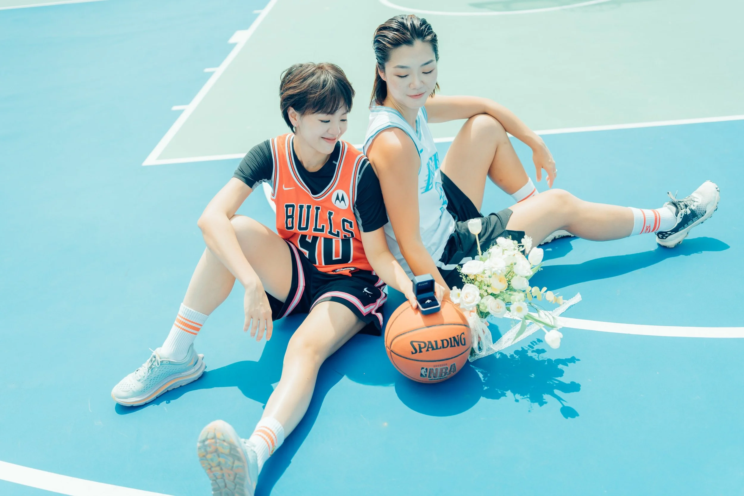 Two women sitting on a blue basketball court, wearing sports jerseys, with a basketball, bouquet of flowers, and a ring box in front of them, celebrating with smiles.