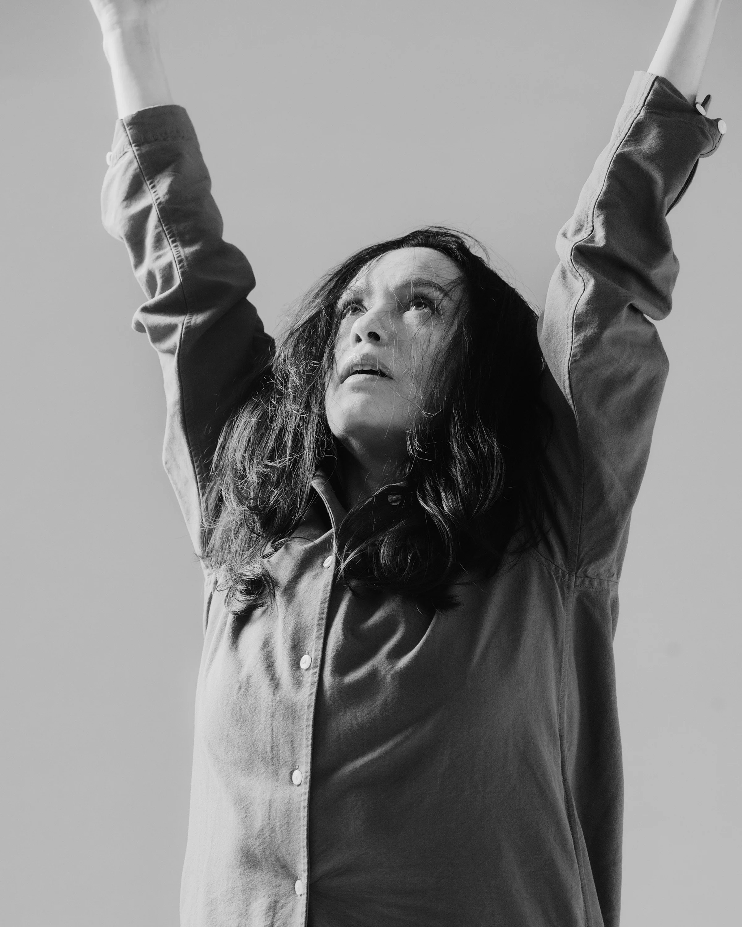 A woman with dark, wavy hair looking upward with raised arms, wearing a button-up shirt, in a black and white photograph.