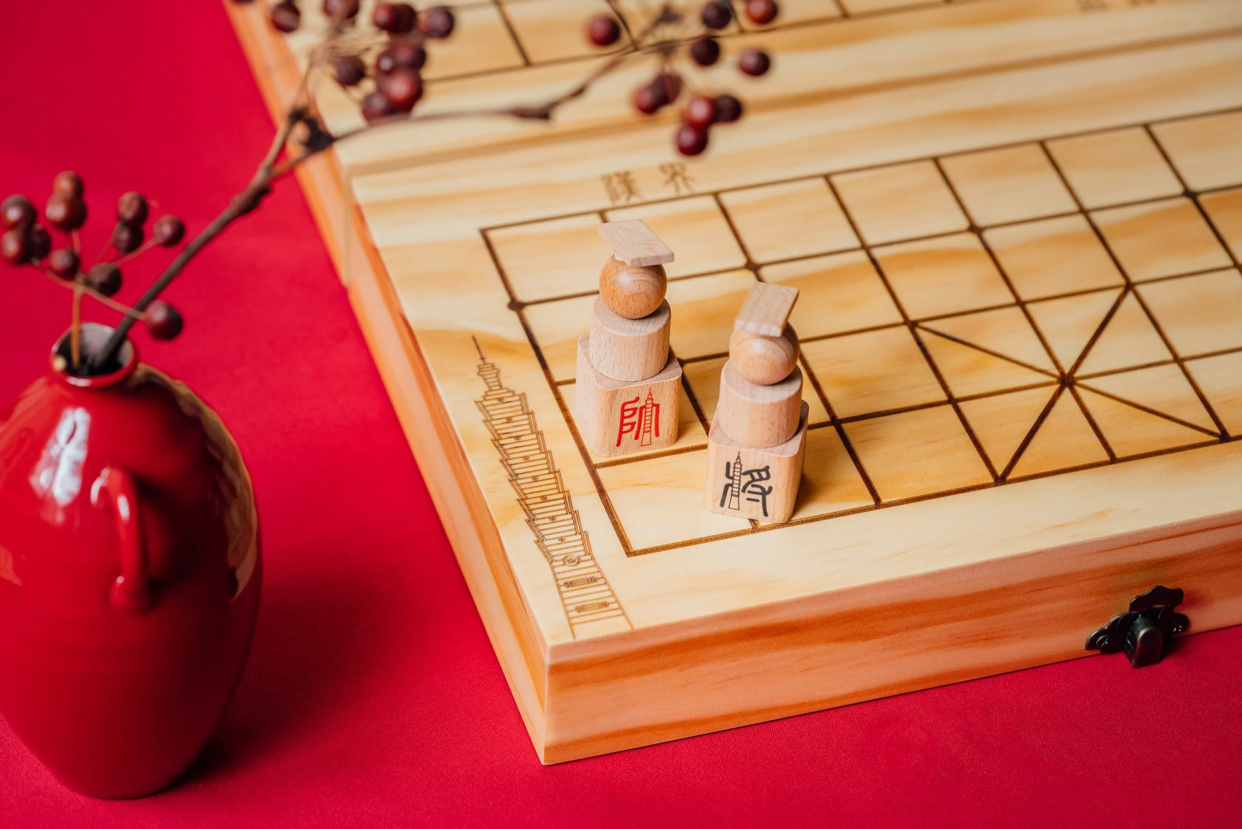 A wooden Chinese chessboard with game pieces and a branch with red berries on a red surface.