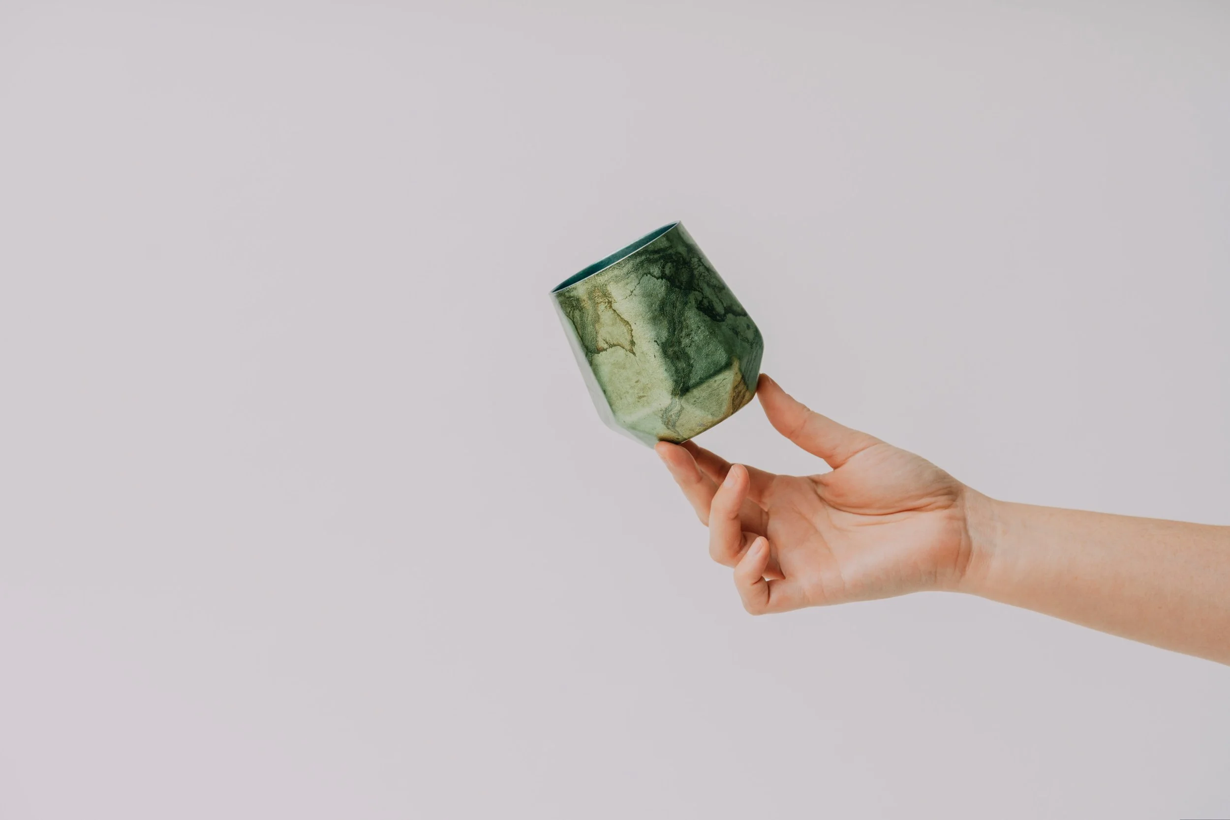 A hand holding a small ceramic cup with a green marble-like pattern against a plain light background.