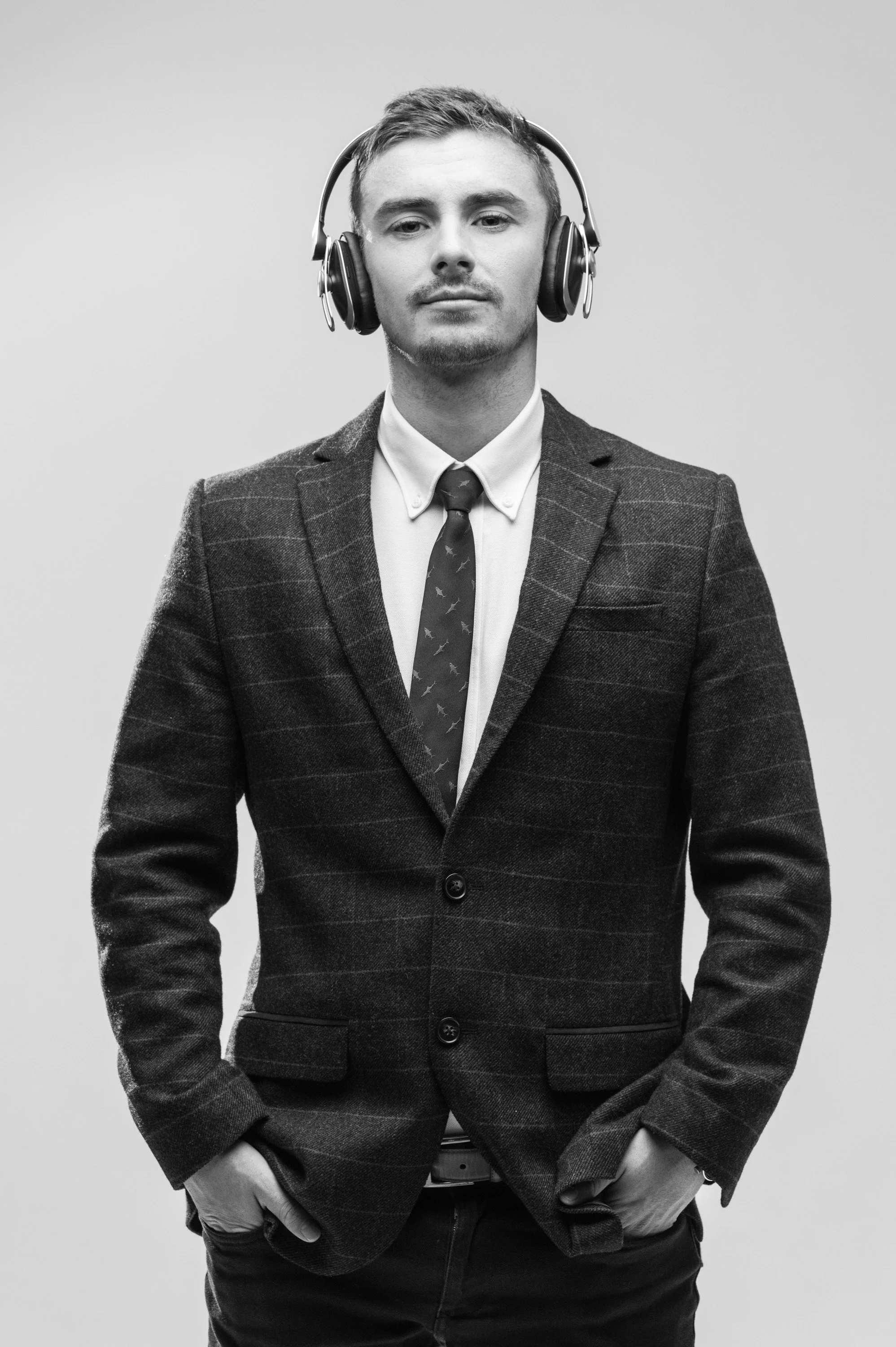 Black and white photo of a man in a business suit and tie, wearing over-ear headphones, standing against a plain background, looking confidently at the camera.