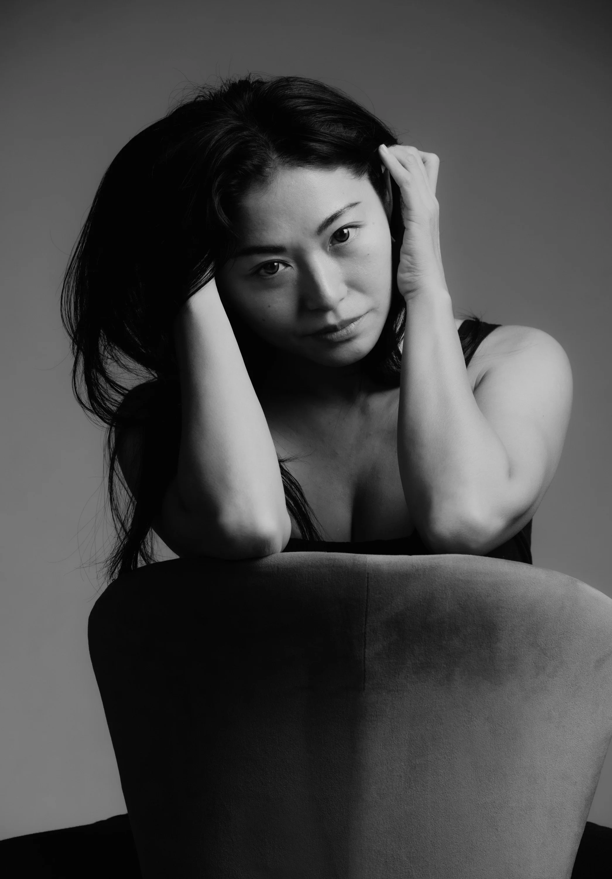 Black and white portrait of a woman with dark hair, looking at the camera, with hands touching her head.