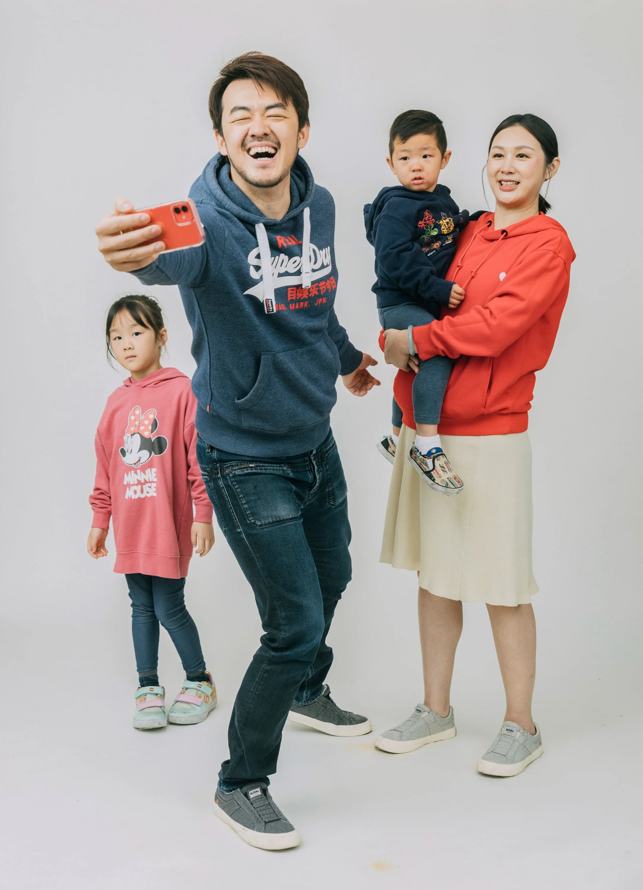 A happy man taking a selfie with a family of four, including a woman holding a young boy and a girl standing nearby, in a studio with a plain white background.