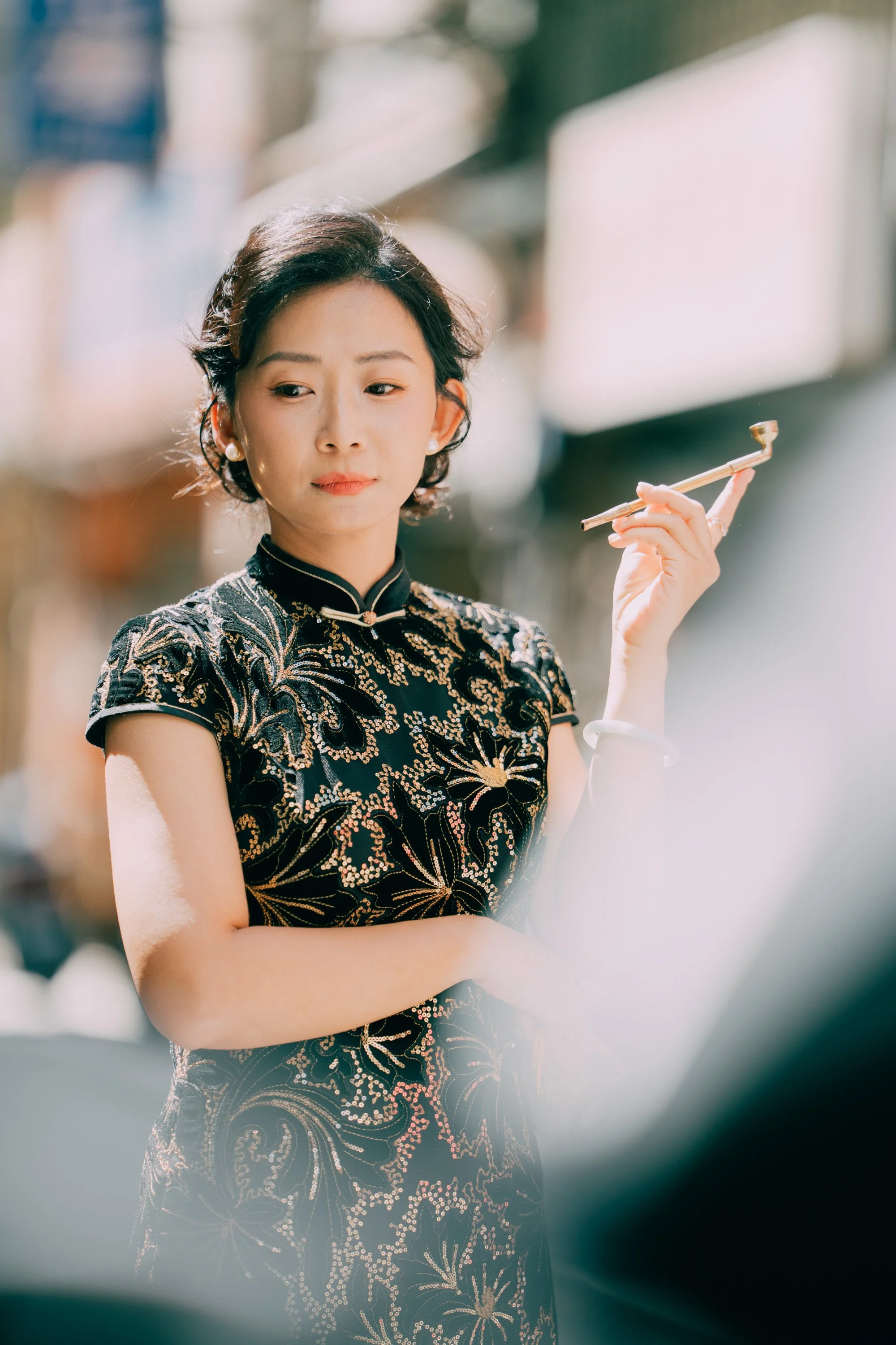A woman with short dark hair and pearl earrings wearing a black dress with gold embroidery, standing outdoors and holding a chopstick in her right hand.