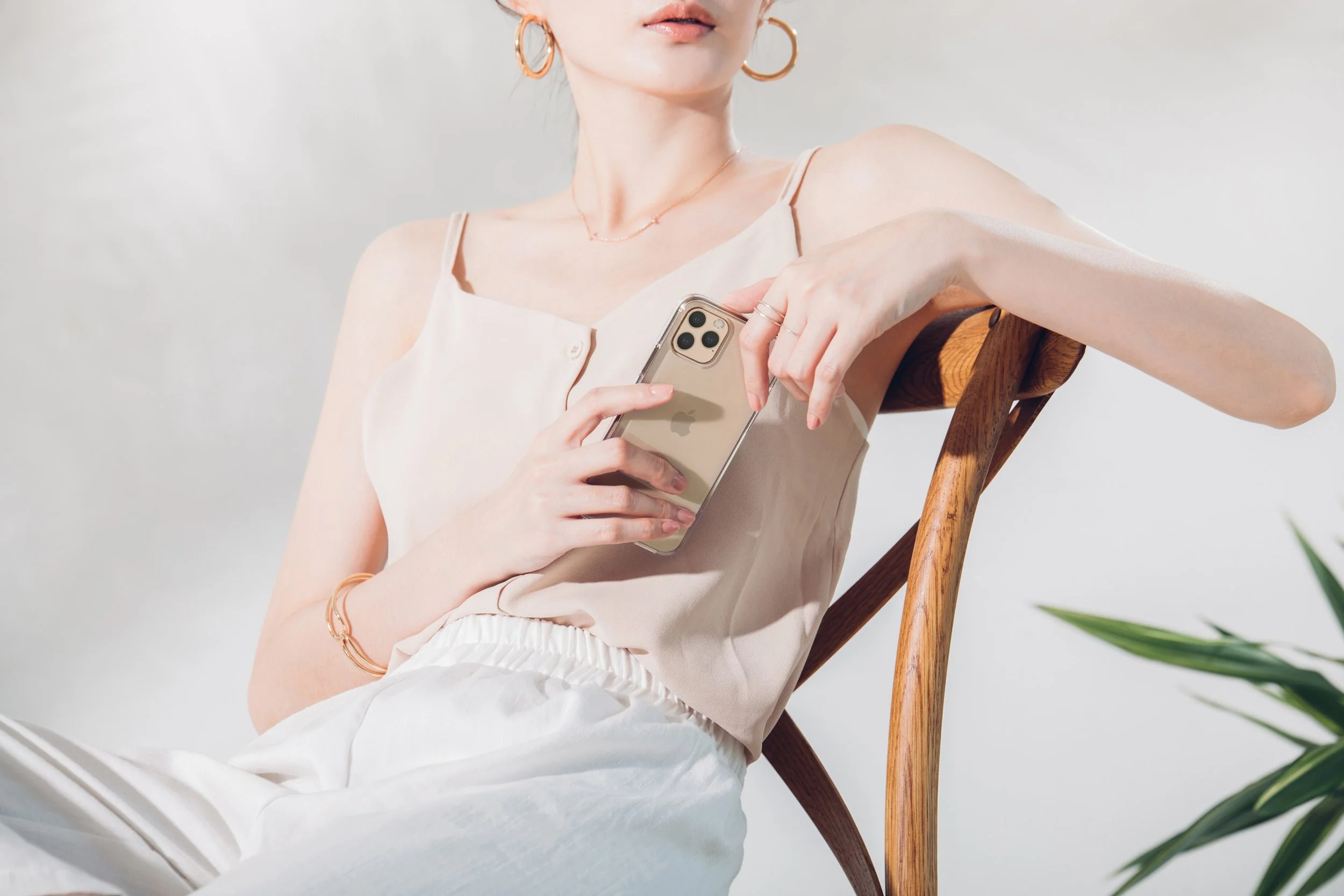 A woman in a light-colored sleeveless top sitting on a wooden chair, holding a smartphone in her hands, with a neutral background and some green plants visible in the corner.
