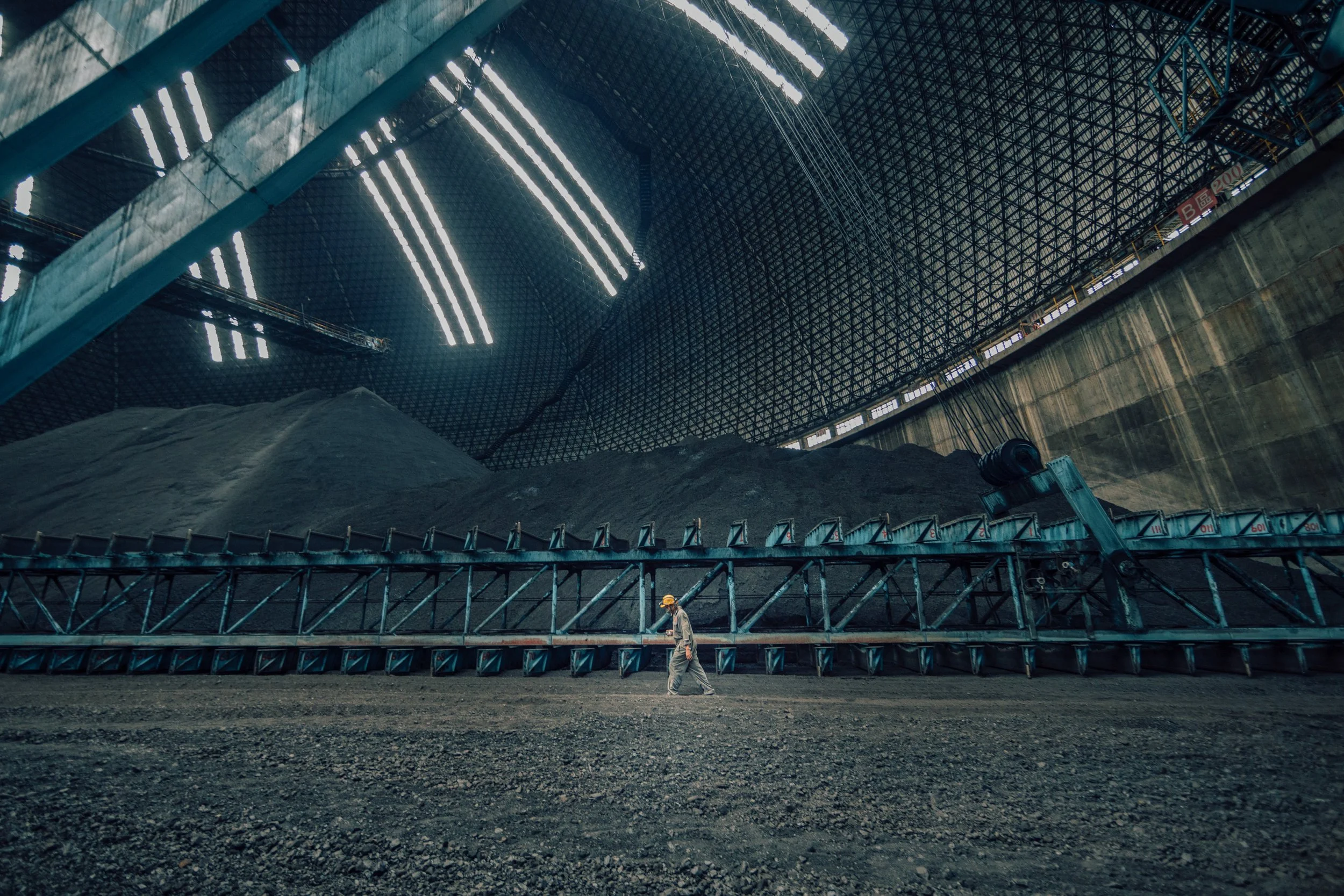 Inside a large industrial building with a high ceiling and a metal grid structure. A worker in a yellow helmet and gray uniform walks on a rough ground surface. There is a conveyor belt with black granules and machinery, with the environment illumina
