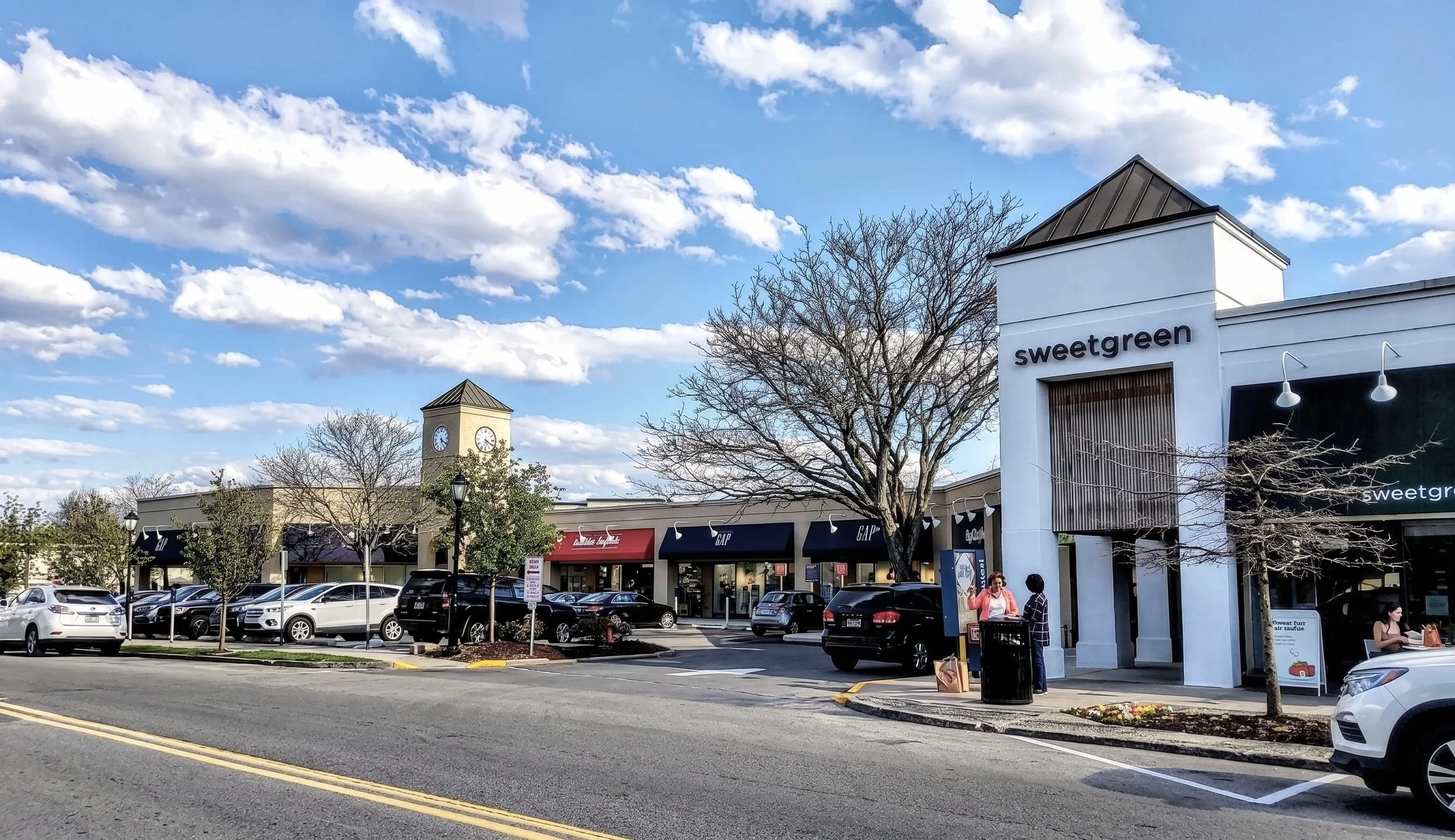 A shopping mall parking lot with cars parked, a clock tower, storefronts including Sweetgreen, and trees with some leaves.