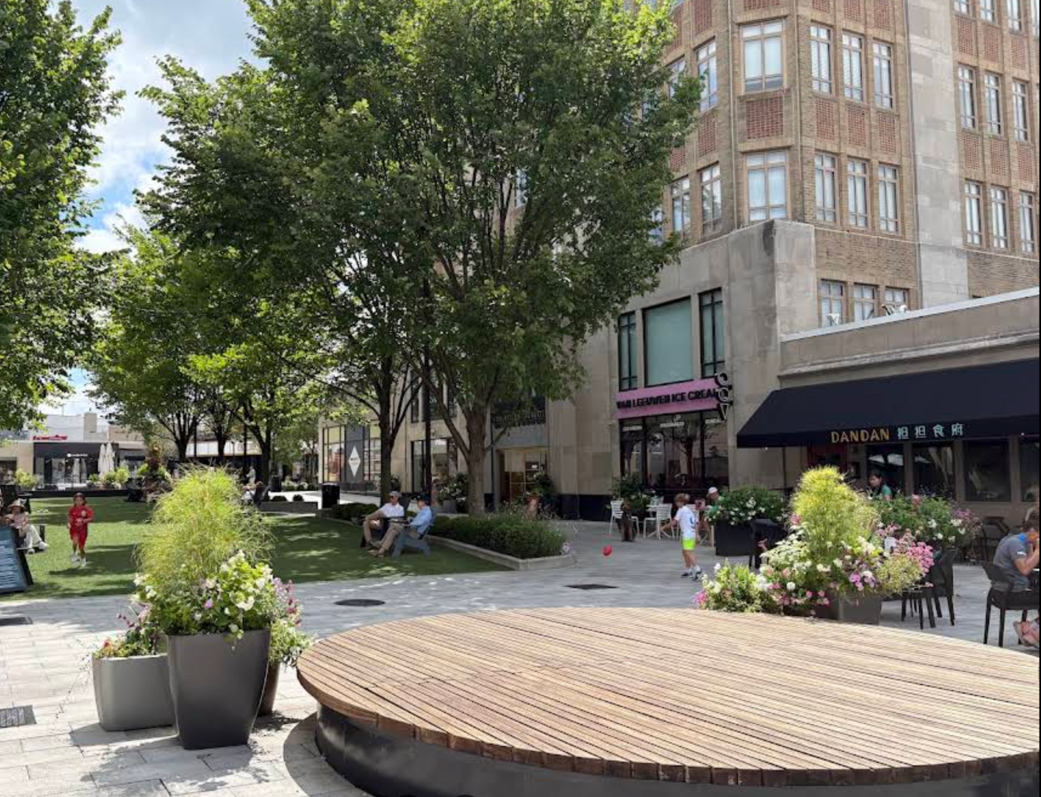 A lively outdoor plaza with people sitting on benches and walking, surrounded by trees, colorful flowers, and a modern building with large windows.