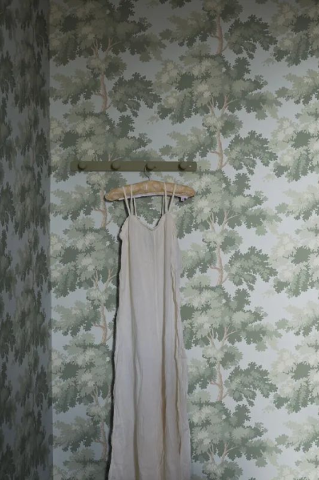 A long white dress hanging on a wooden hanger on a wall-mounted hook in front of a wallpapered wall with a green leaf pattern.
