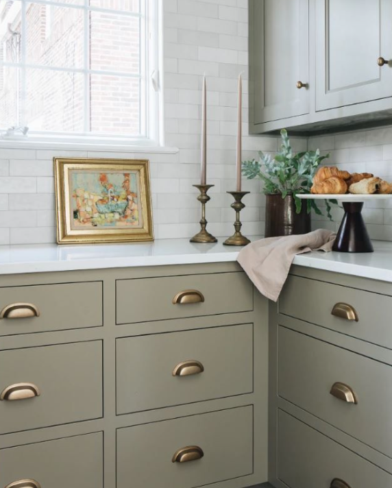 Kitchen corner with light green cabinets, a white countertop, a window, and decor including a framed picture, two tall candlesticks, a potted plant, and a plate with bread.