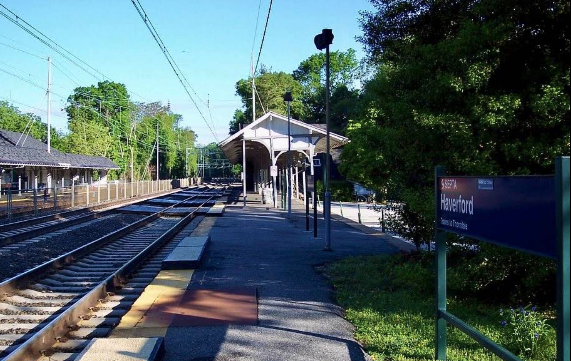 Havertord train station platform with a covered shelter, train tracks running alongside, trees in the background, and a sign reading 'Havertord'.