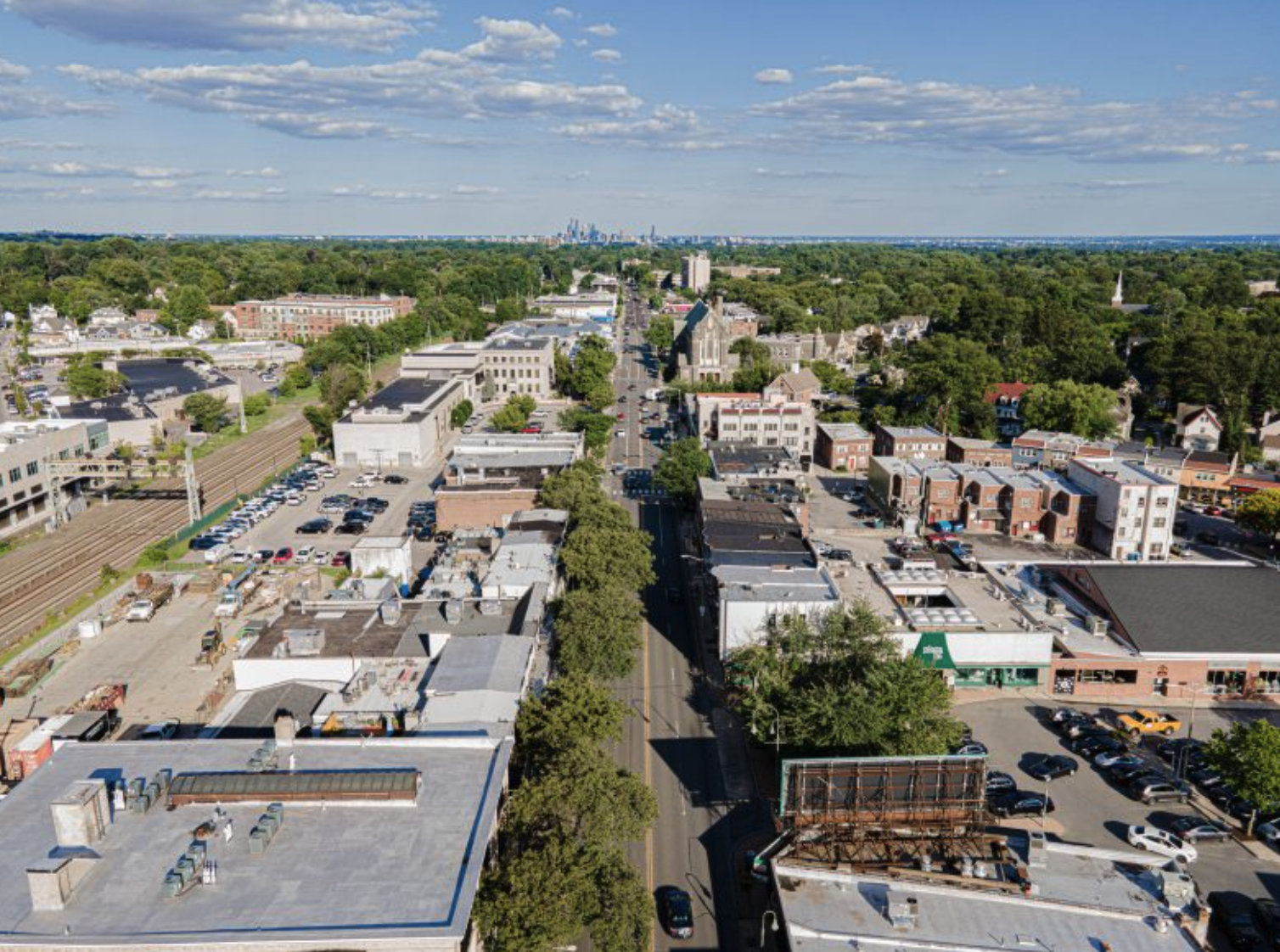 Aerial view of a city street with buildings, trees, parked cars, and a train track on the left side under a partly cloudy sky.