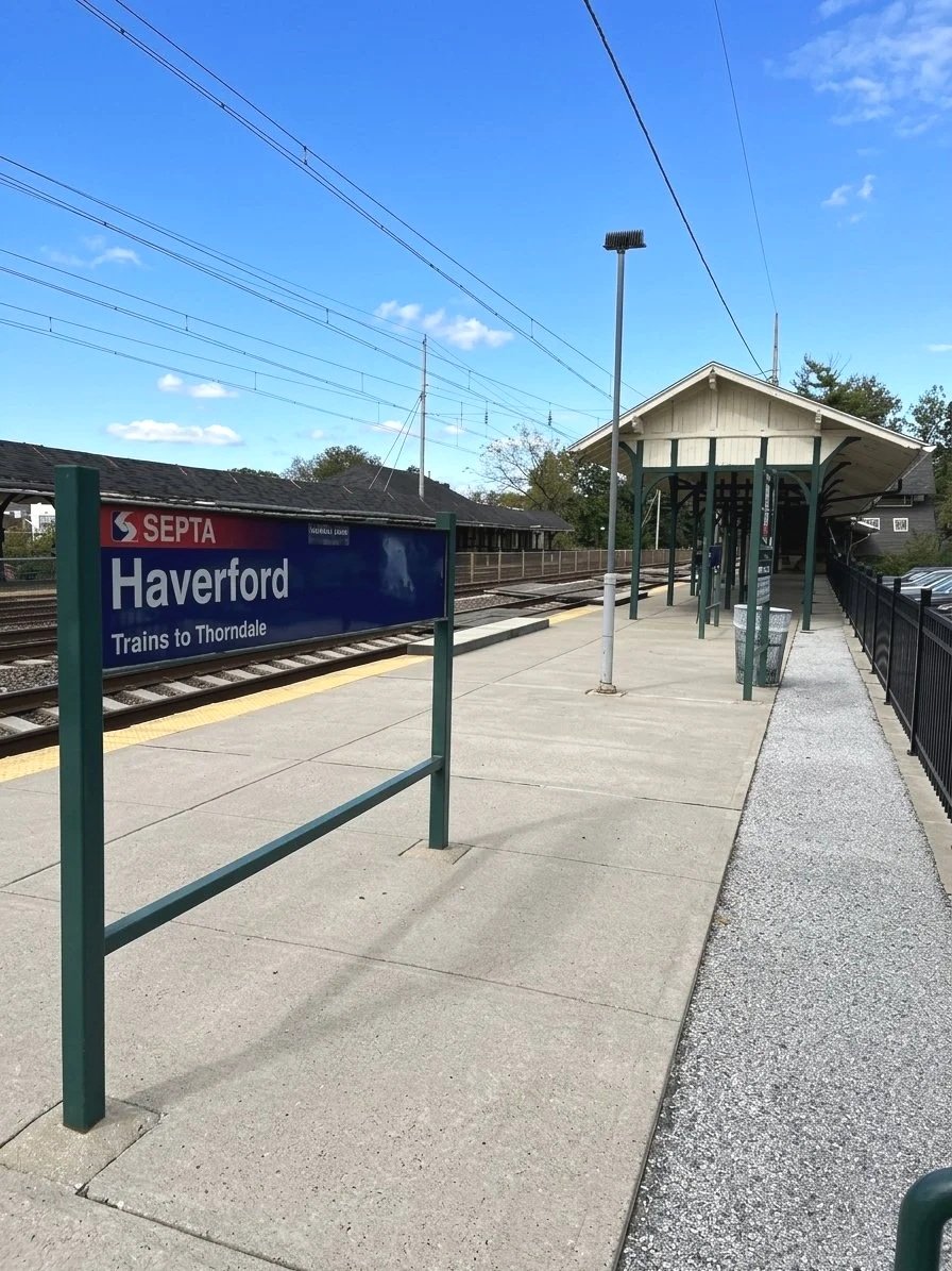 A train station platform at Haverford with a sign indicating trains to Thorndale, a shelter, a light pole, and overhead train tracks under a partly cloudy blue sky.
