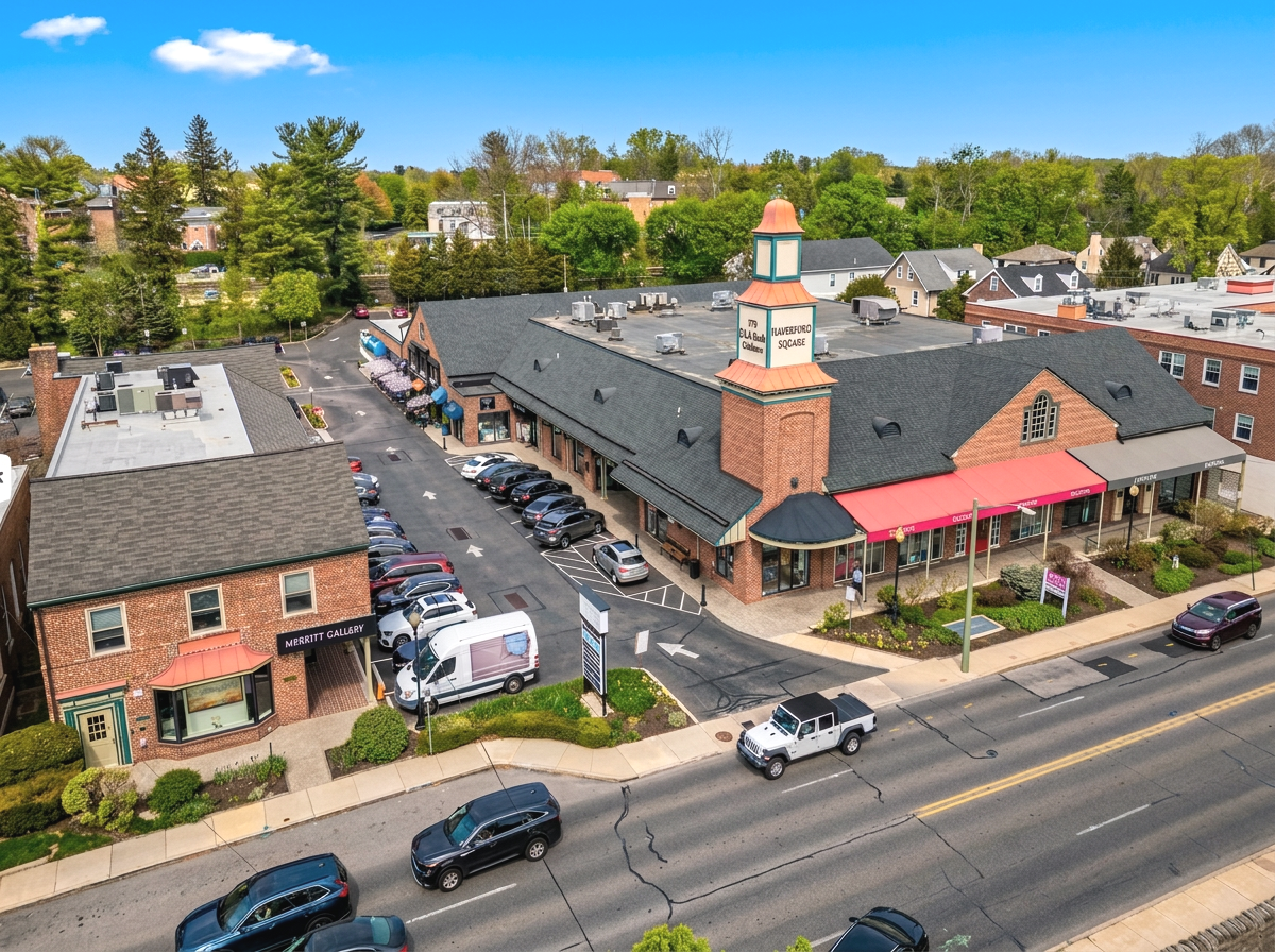 An aerial view of a shopping center with parking lots, cars, and buildings in a small town surrounded by green trees and houses under a partly cloudy sky.