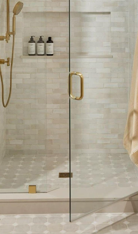 A modern glass shower with a gold door handle and a rainfall showerhead, white tiled walls, built-in shelves with black bottles, and a tiled floor.