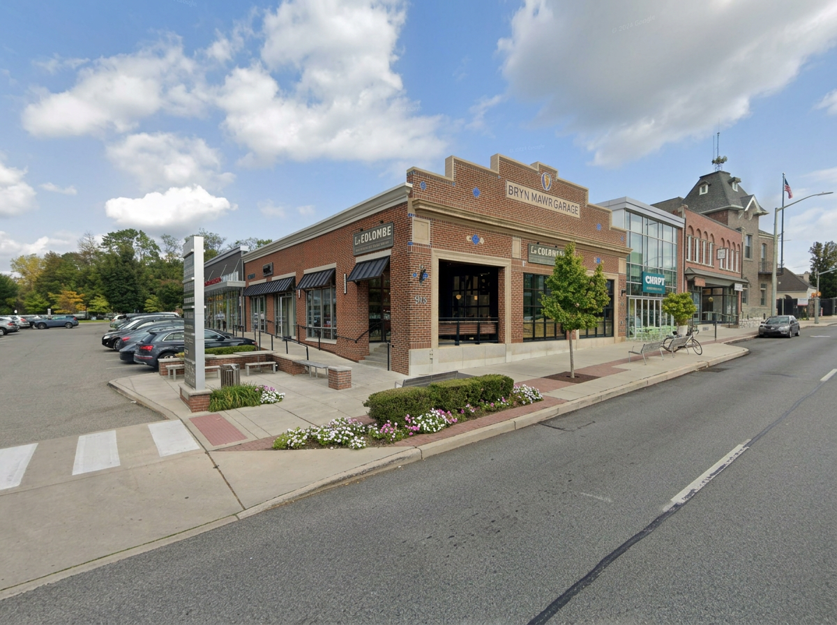 A brick commercial building with name signs and shops, including Bryn Mawr Garage, in a small town with parked cars, a sidewalk, small trees, and a church in the background under a partly cloudy sky.