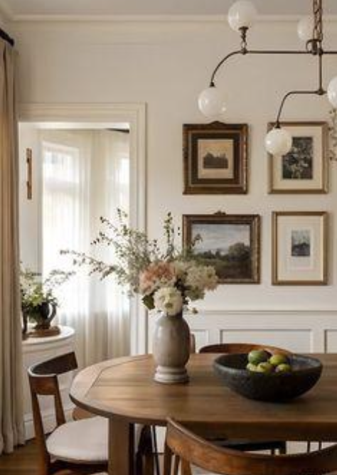 A round wooden dining table with a vase of flowers and a bowl of apples, surrounded by wooden chairs in a room with framed artwork on the wall and light streaming through a window.