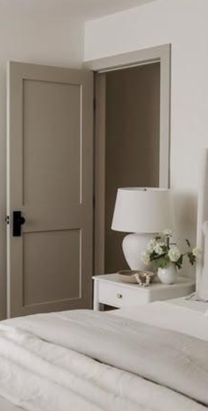 A bedroom with a partially open door, a bedside table with a white lamp and a flower arrangement, and a neatly made bed with white linens.