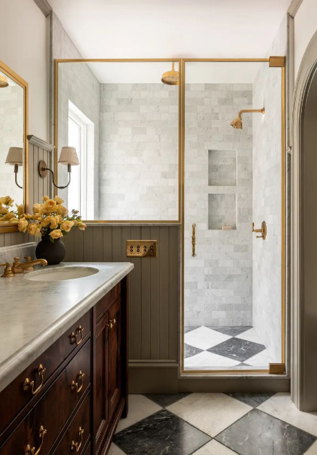 Bathroom with a marble countertop vanity, a mirror, wall-mounted light fixtures, a vase with yellow flowers, a glass shower enclosure with gold trim, shower niche, checkered black and white floor tiles, and a window.
