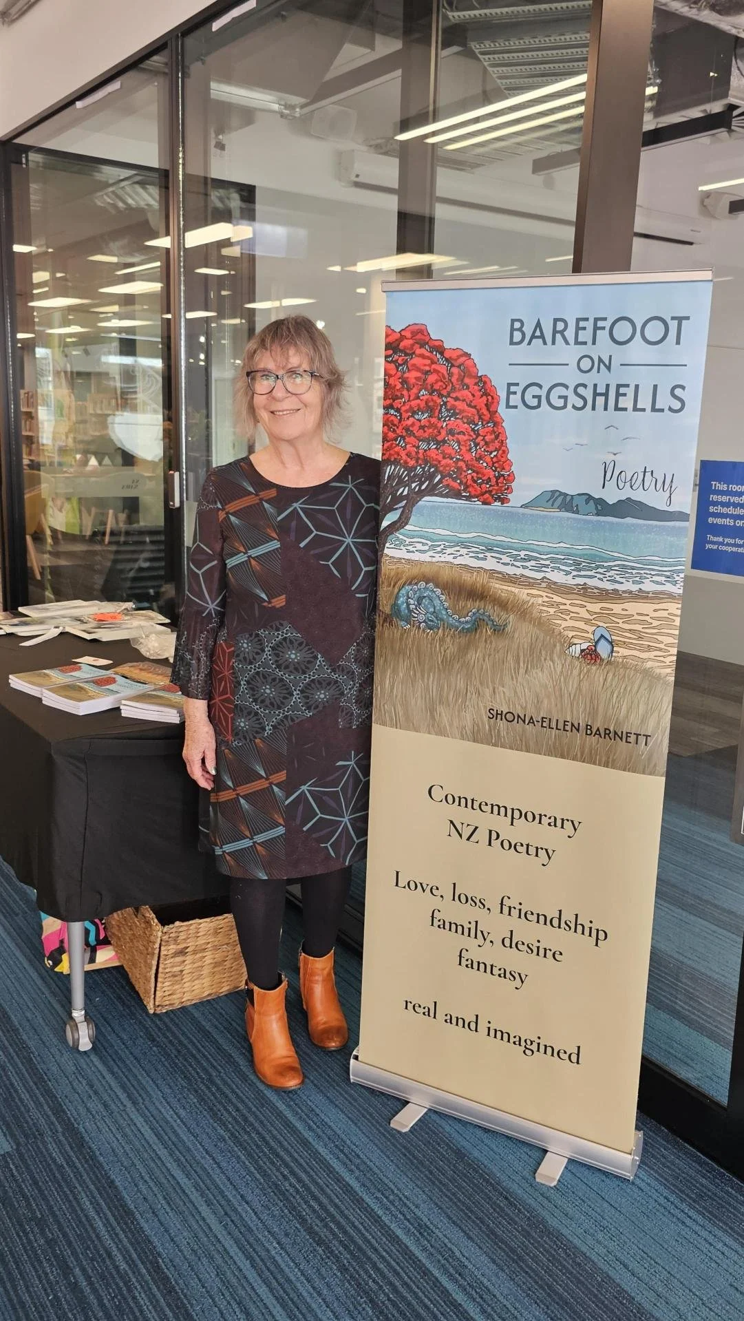 A woman standing next to a tall, vertical banner promoting a book titled 'Barefoot on Eggshells' by Shona-Ellen Barnett. The banner features artwork of a red-leaved tree, ocean, and grass, with text describing contemporary NZ poetry about love, loss, friendship, family, desire, fantasy, and real and imagined worlds. The woman is wearing an abstract patterned dress, black tights, and orange boots, smiling at the camera in an indoor setting.