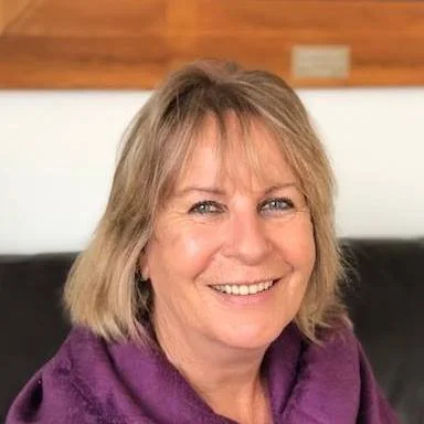 A smiling woman with blonde hair, wearing a purple top, sitting on a black couch with a wooden wall background.