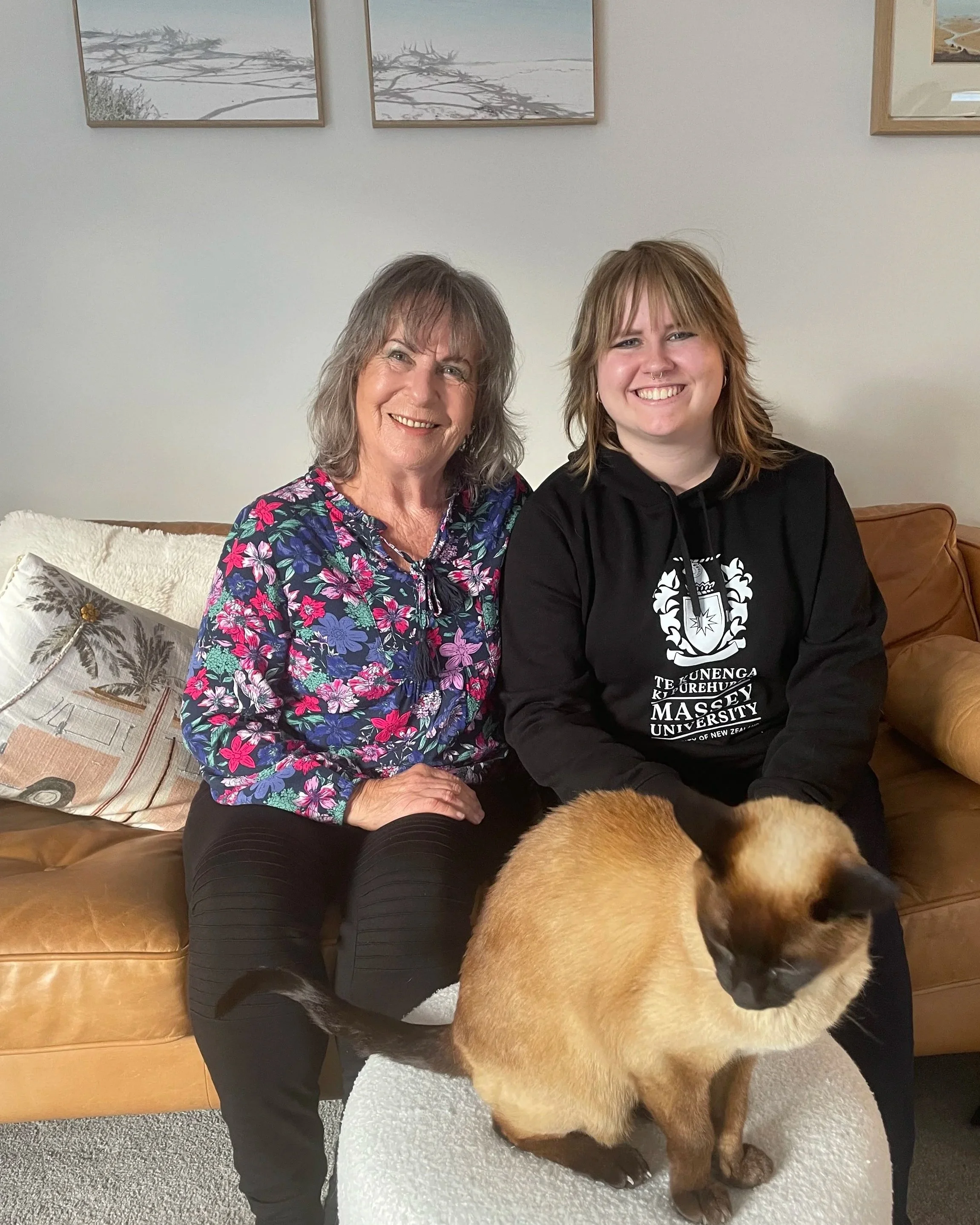 Two women sitting on a couch, smiling, with a large dog in front of them. The woman on the left has shoulder-length gray hair and is wearing a colorful floral blouse. The woman on the right has shoulder-length light brown hair with bangs and is wearing a black hoodie with white text and graphics. The dog is a large, tan-colored breed with a dark face, sitting on a white fluffy bed. The background features framed artwork on a white wall.