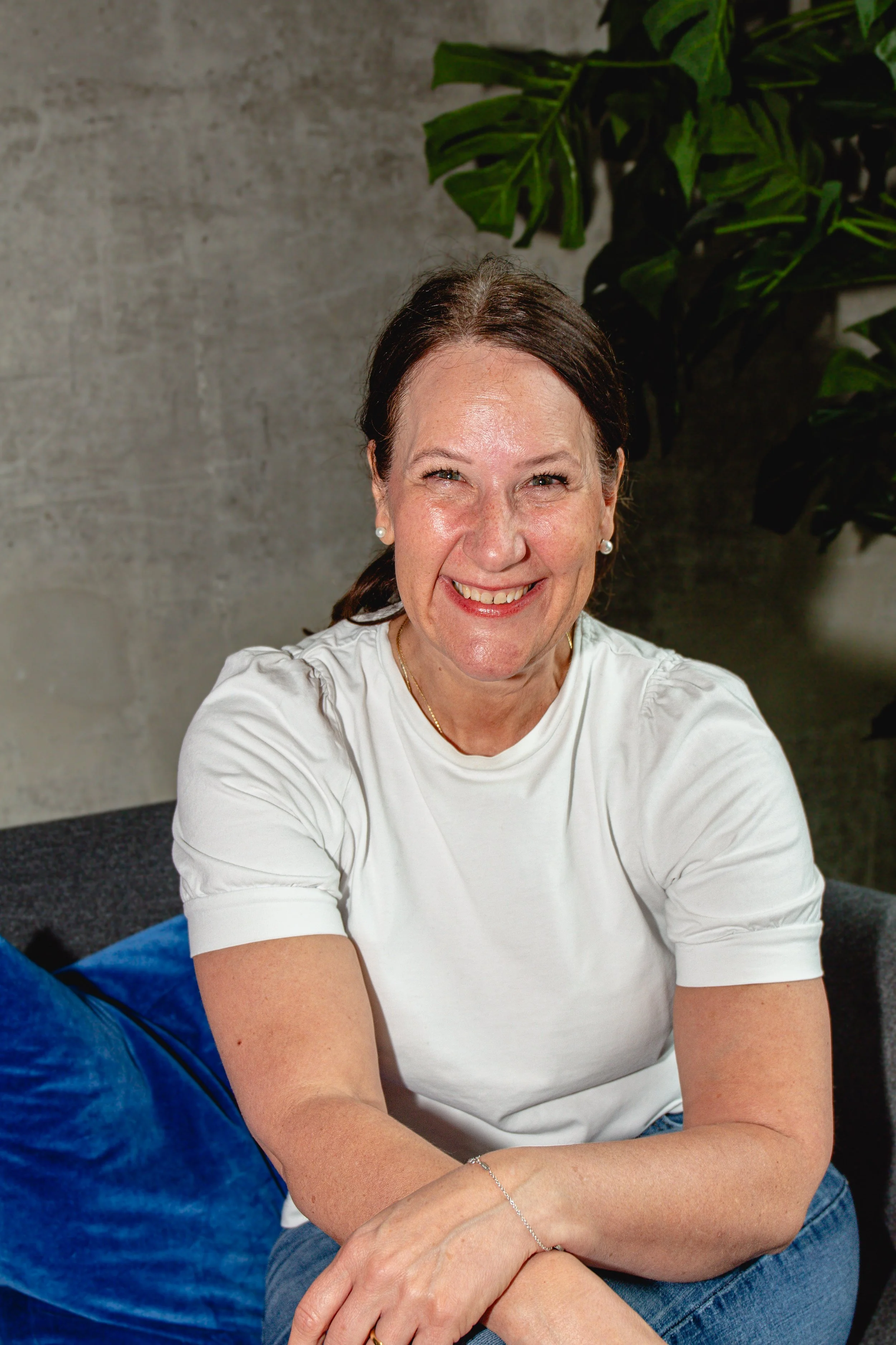 A woman with reddish-brown hair, smiling, wearing a light blue sweater, sitting on a beige couch with a pillow behind her, in a bright room with white curtains and a potted plant.
