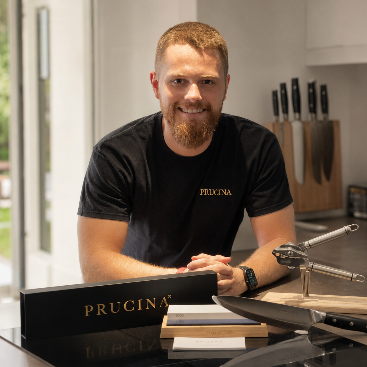 A smiling man with a red beard and short hair, wearing a black T-shirt with the word PRUCINA, sitting at a kitchen counter with knives in the background and various kitchen tools and sharpening stones in front of him.