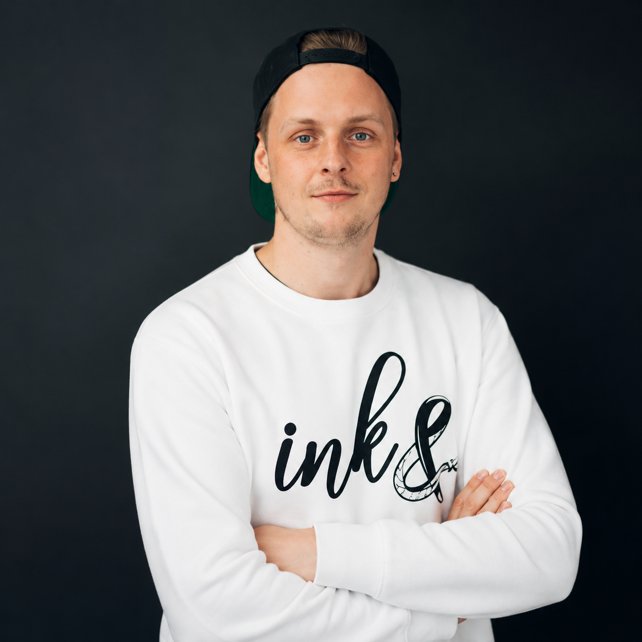 A young man with light skin, blue eyes, and light brown hair is wearing a black backward cap and a white sweatshirt with black text that says 'ink &', with a tattooed feather illustration. He has a slight smile and is standing with his arms crossed against a dark background.