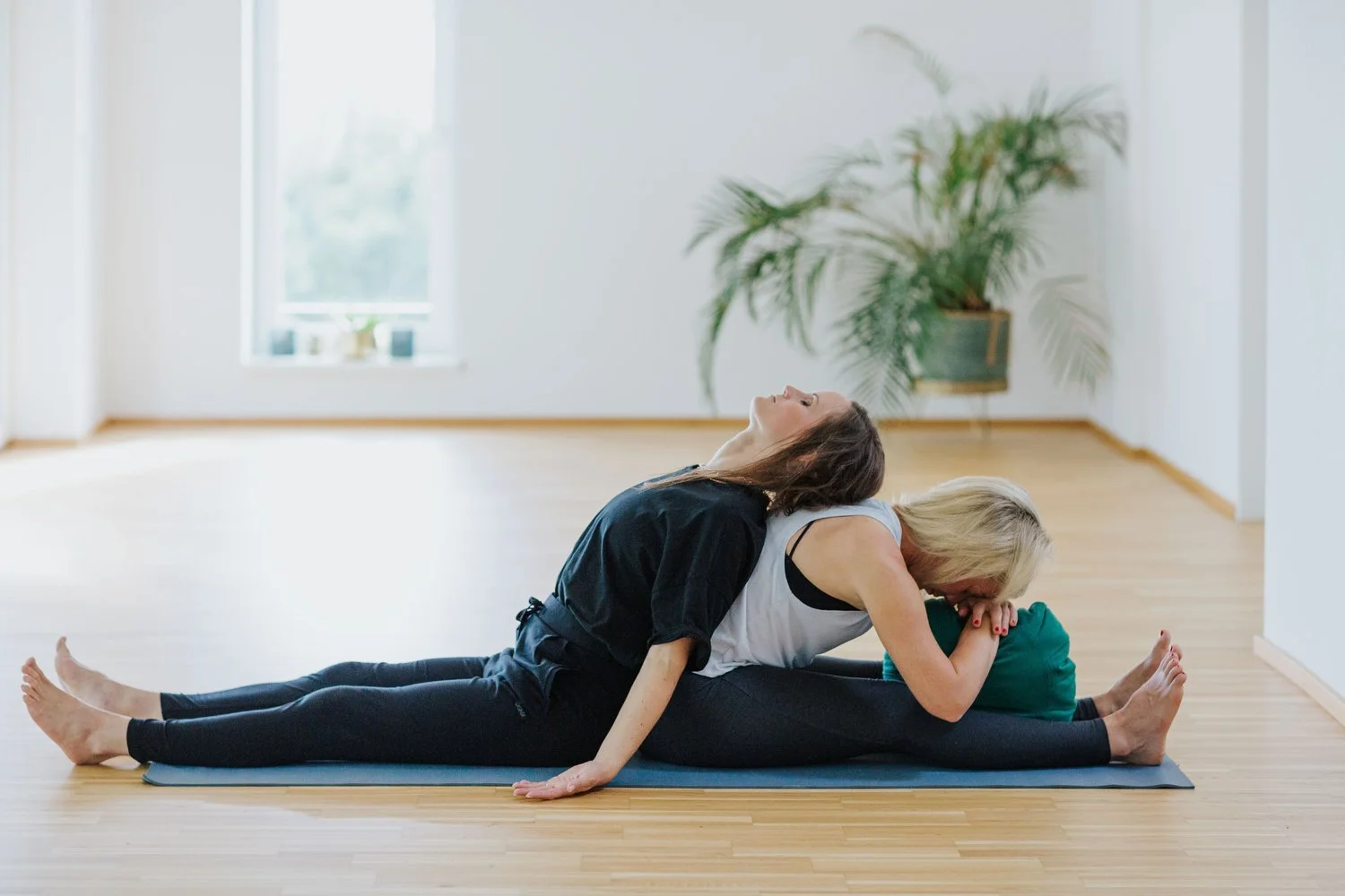 Zwei Frauen in einer Yoga-Pose in einem hellen Raum mit Parkettboden und Pflanzen.