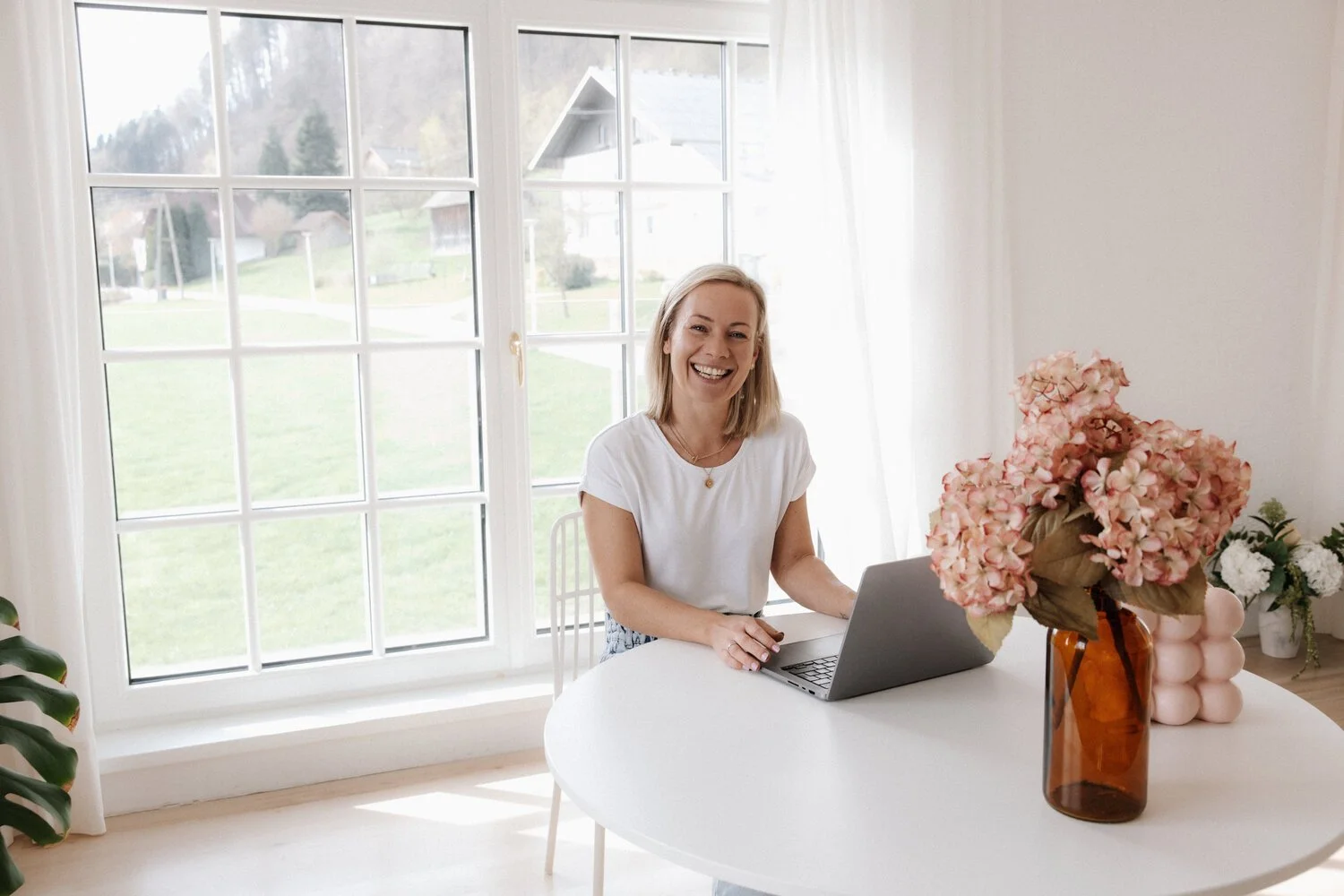 Eine Frau sitzt an einem weißen Tisch mit einem Laptop, umgeben von Blumenvasen mit Blumen in einem hellen Raum mit großen Fenstern und Blick ins Grüne.
